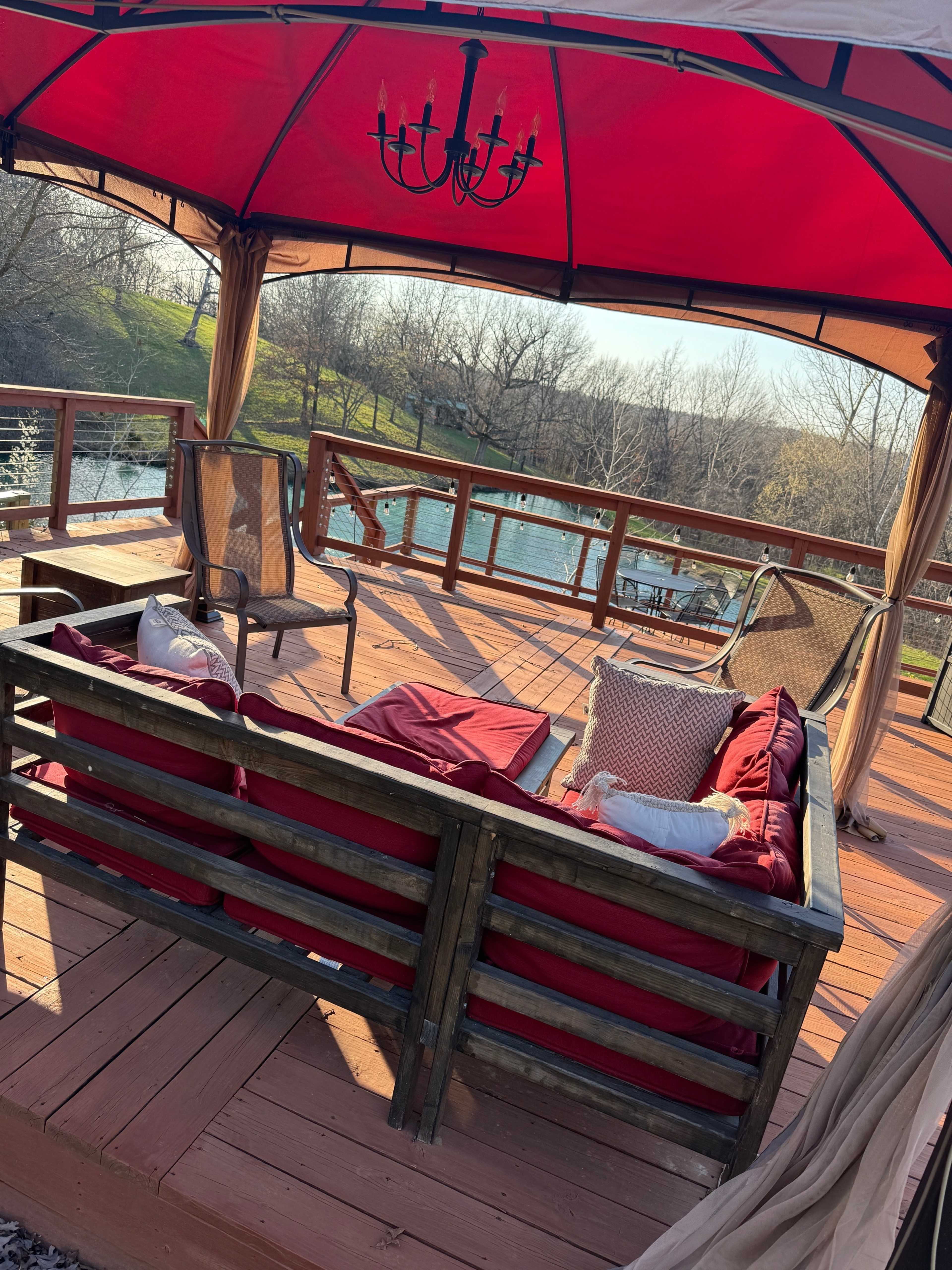 A wooden patio features a canopy with a chandelier, surrounding a seating area overlooking a body of water and trees in the background.