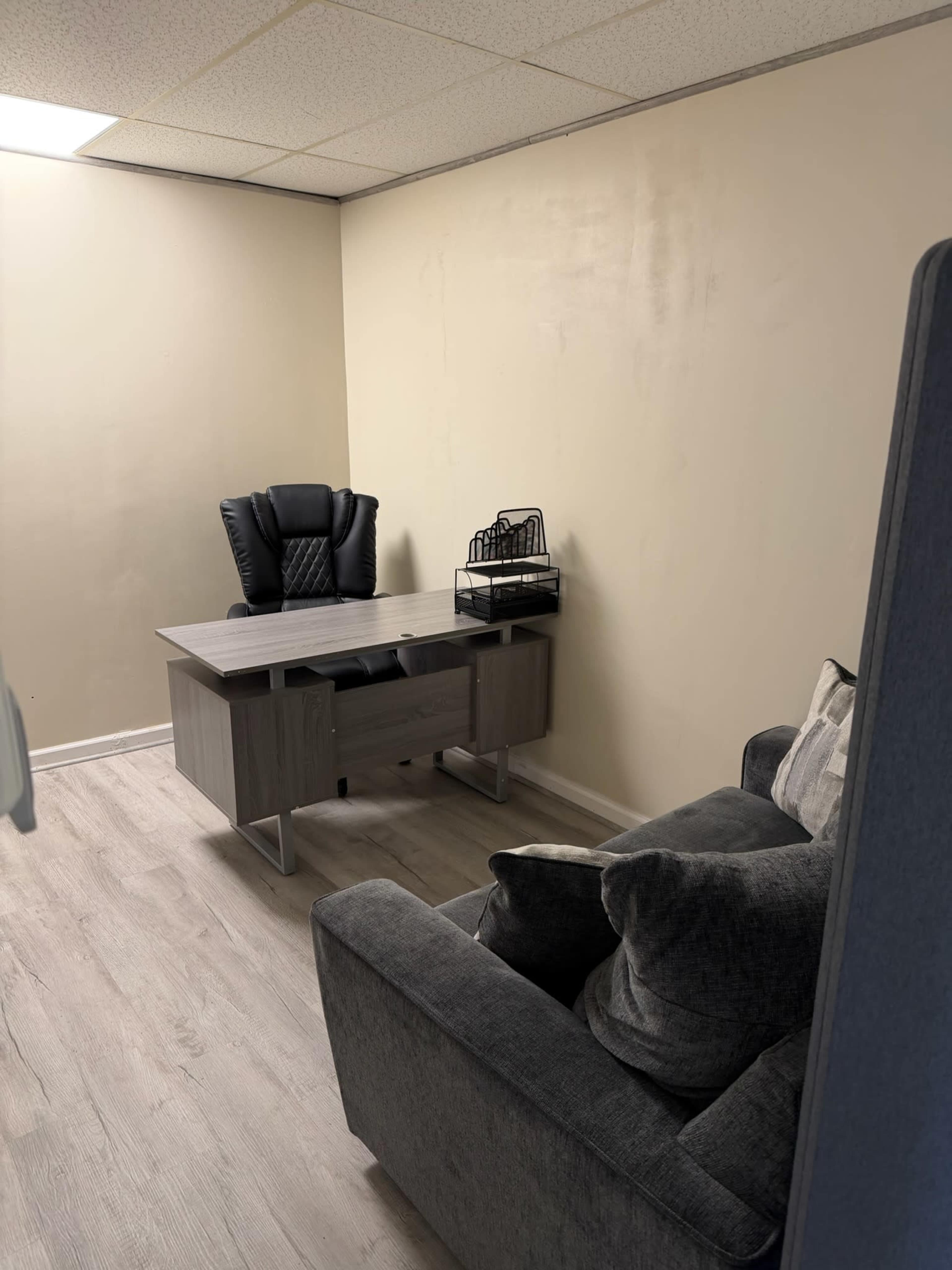The image shows an office space featuring a gray sofa, a modern desk with a chair, and a small decorative item on the desk against a pale wall.