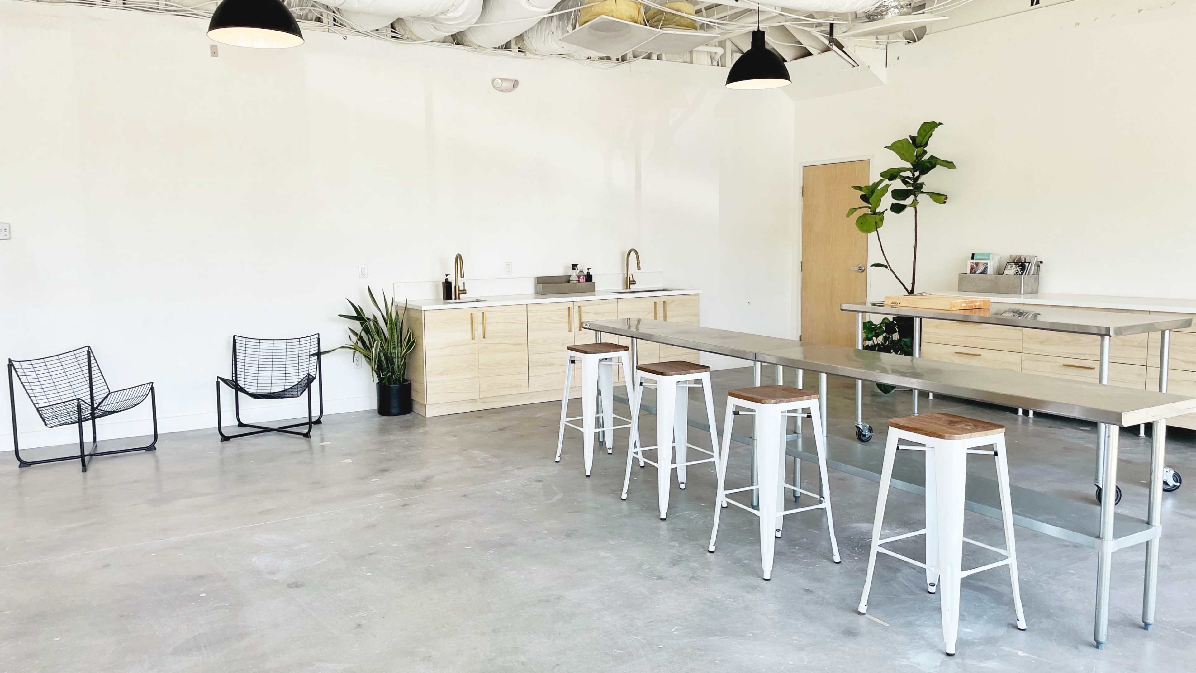 A spacious room features a kitchen area with wooden cabinetry, a metal table with stools, and two wire chairs, along with a plant in the corner.
