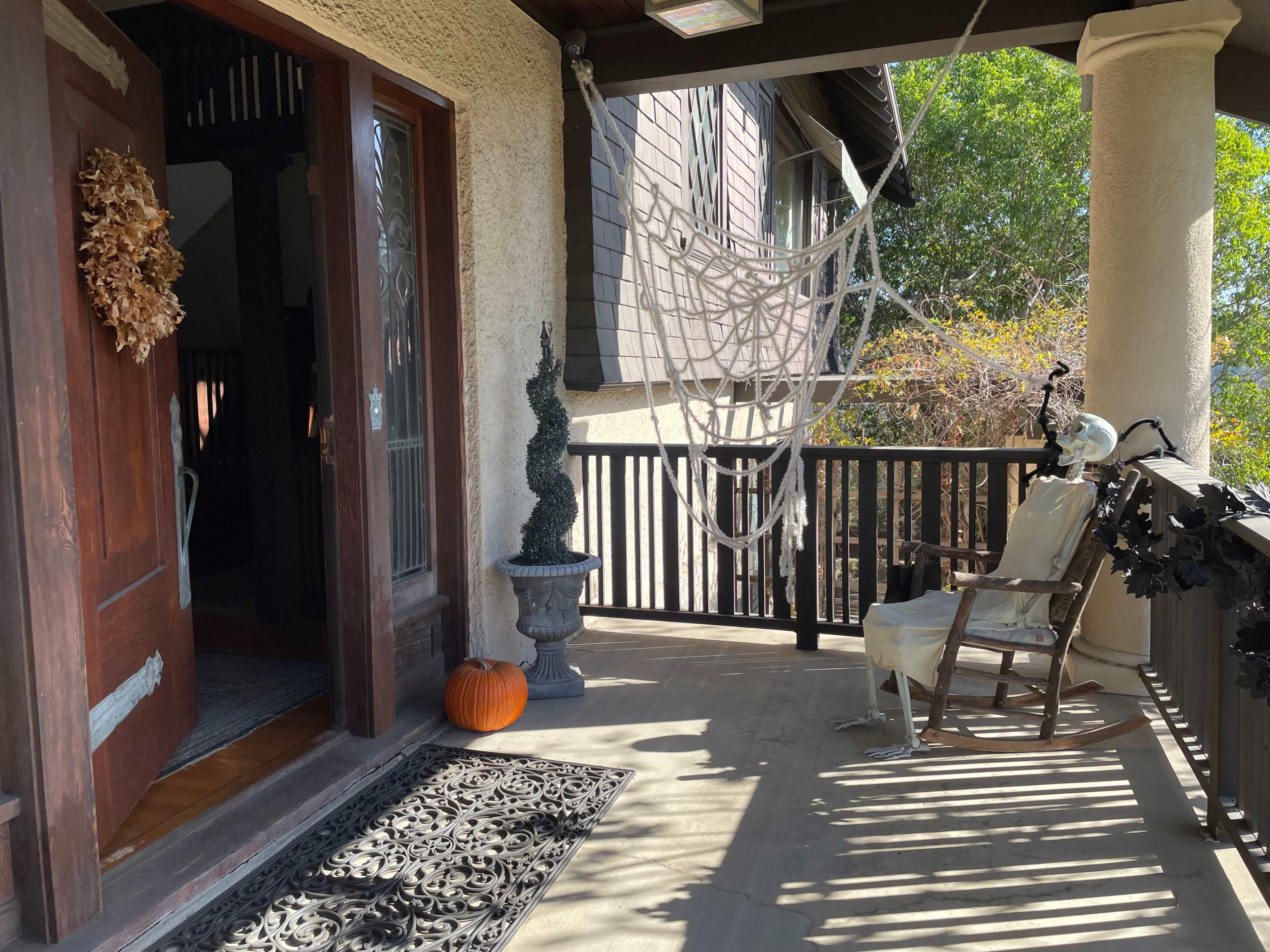 A skeleton dressed in a white cloak sits in a rocking chair on a porch decorated with a spider web and an orange pumpkin.