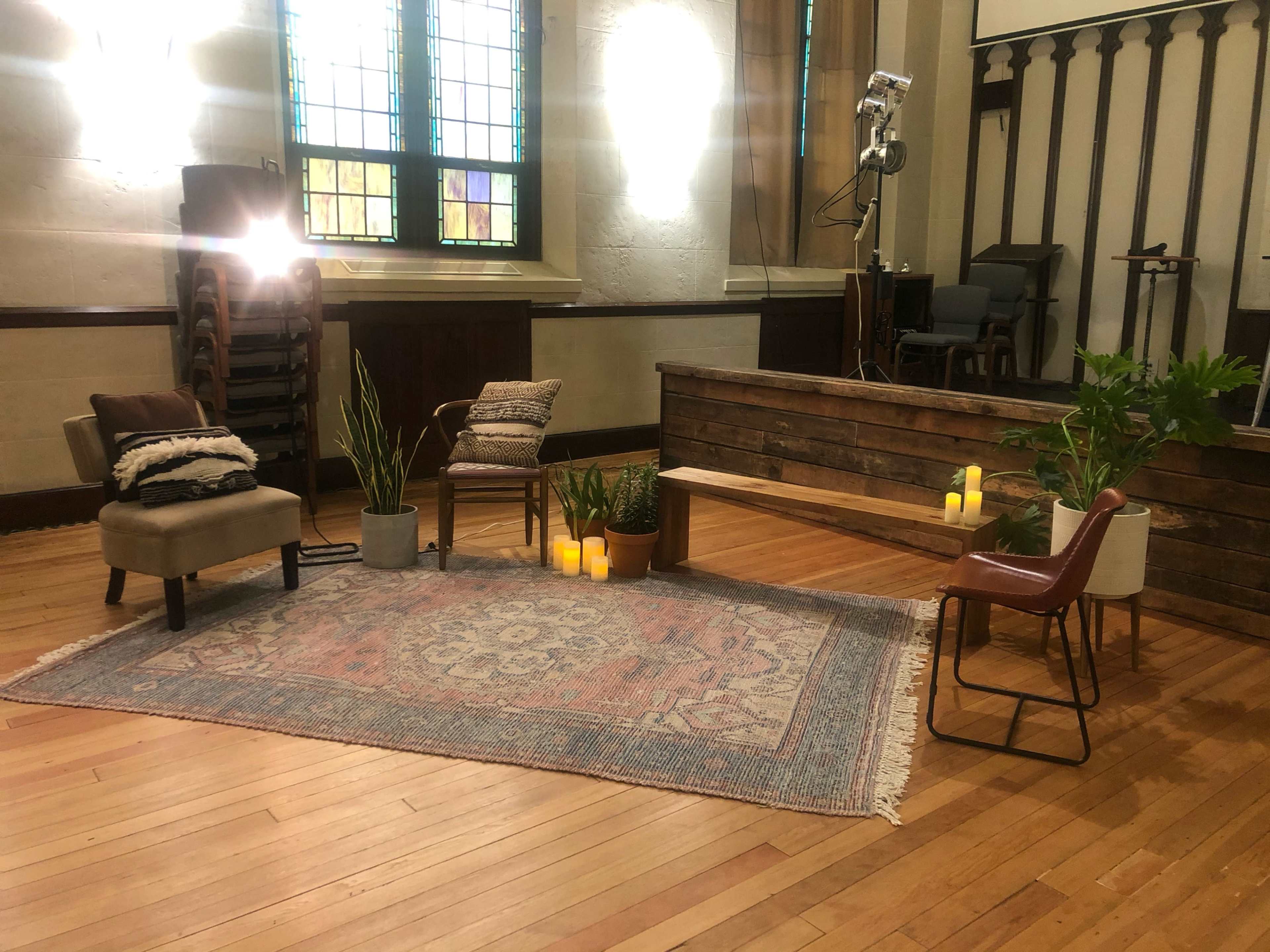 A cozy interior space with a wooden bench, two chairs, potted plants, a patterned rug, and candles arranged in a welcoming layout.