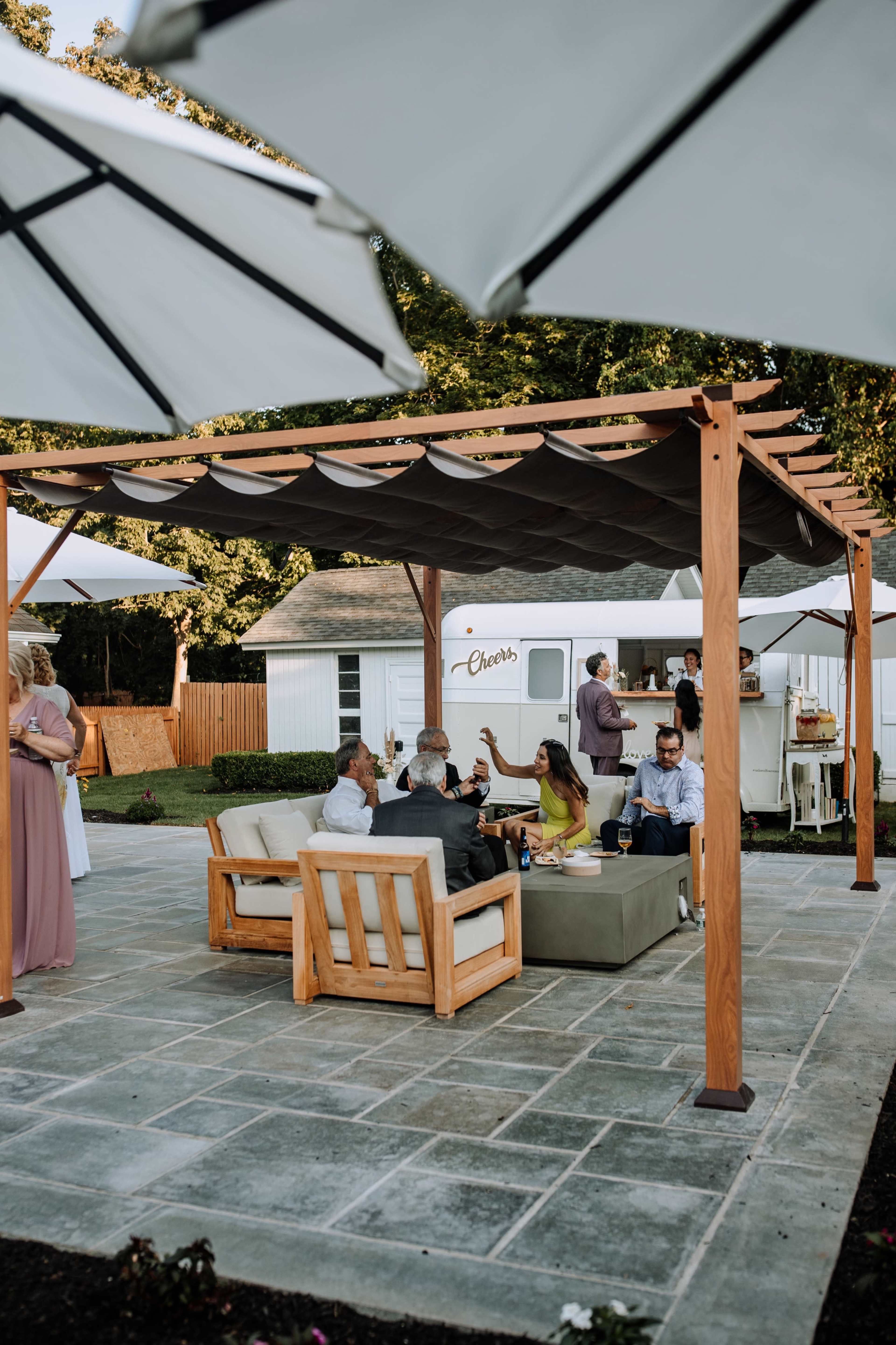 A group of people socialize under a wooden pergola with a shaded seating area at an outdoor event.