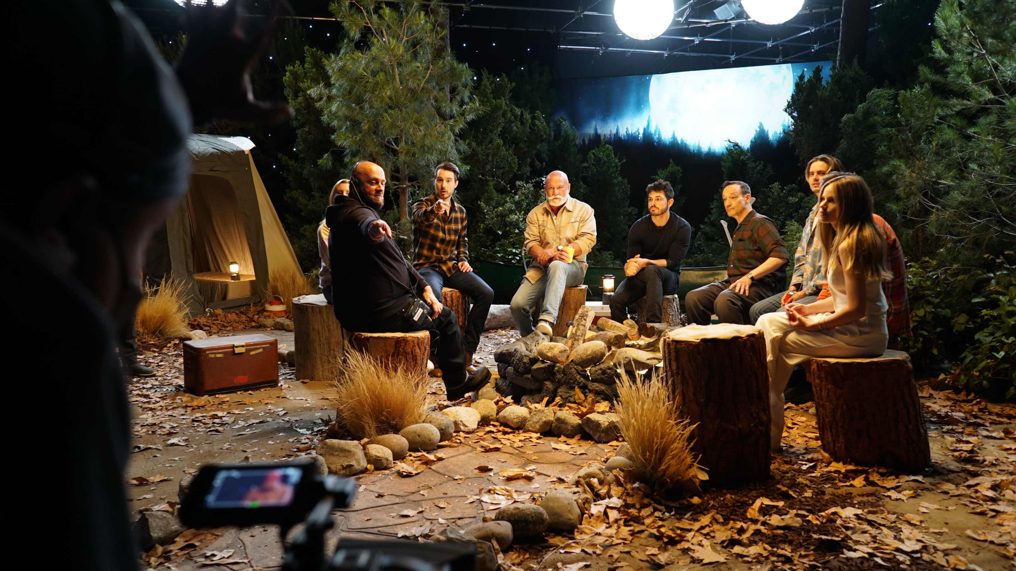 A group of eight people sits around a campfire in a forested studio setting, with a tent and trees in the background.