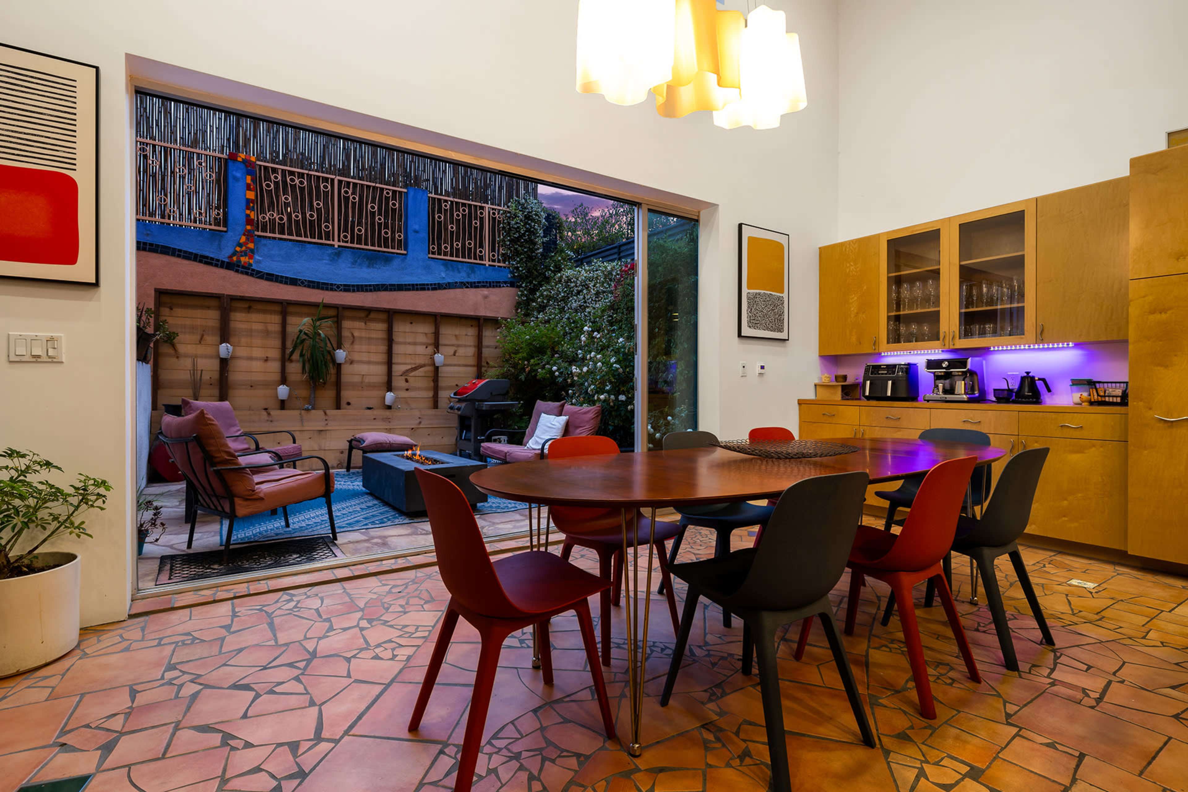 A dining area features a circular wooden table surrounded by colorful chairs, with large sliding glass doors opening to a garden space filled with outdoor furniture and plants.