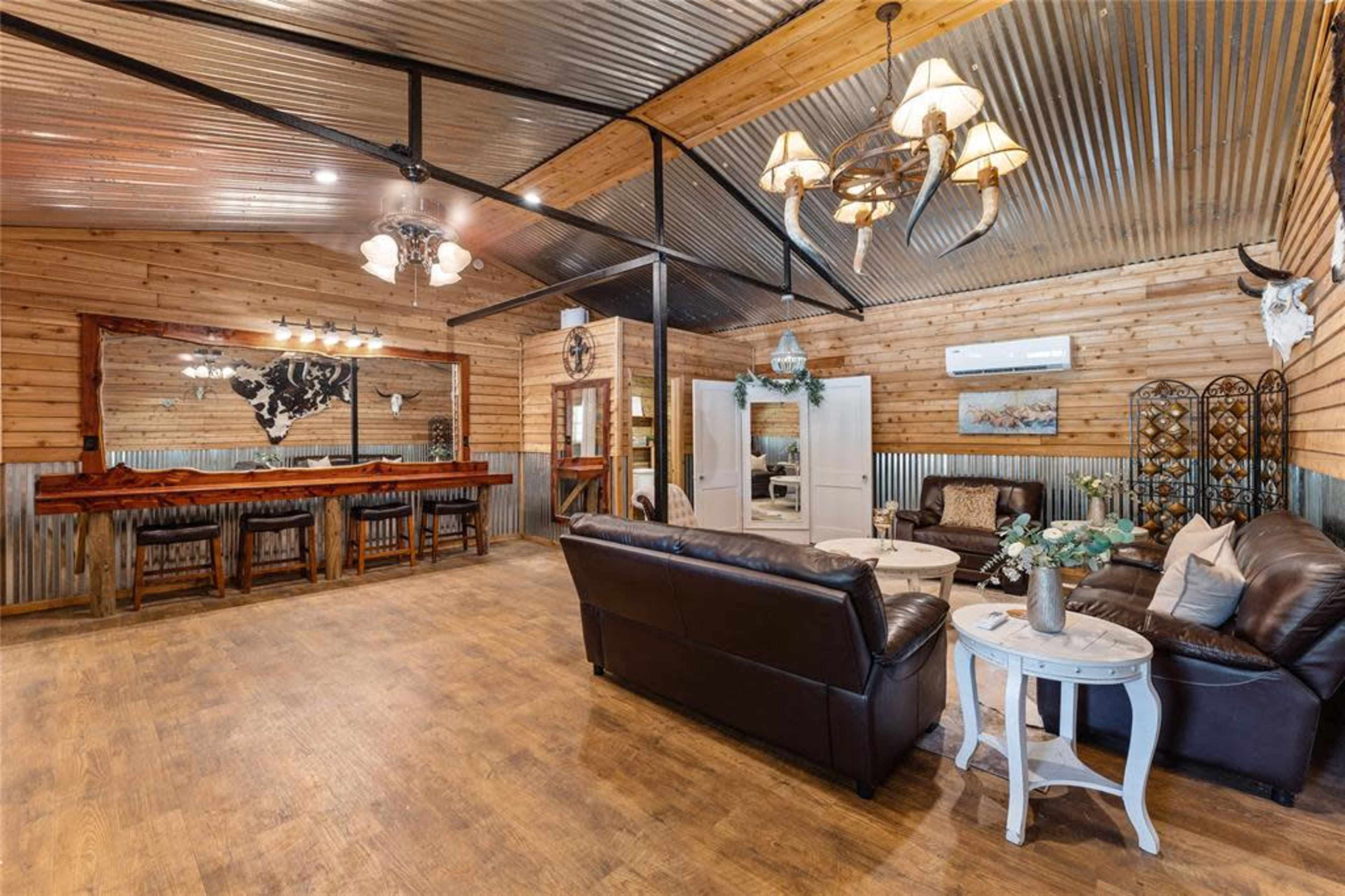 The image showcases a rustic living space featuring leather sofas, a wooden bar area with high chairs, and decorative wall elements, all enclosed by wood-paneled walls and a metal roof.