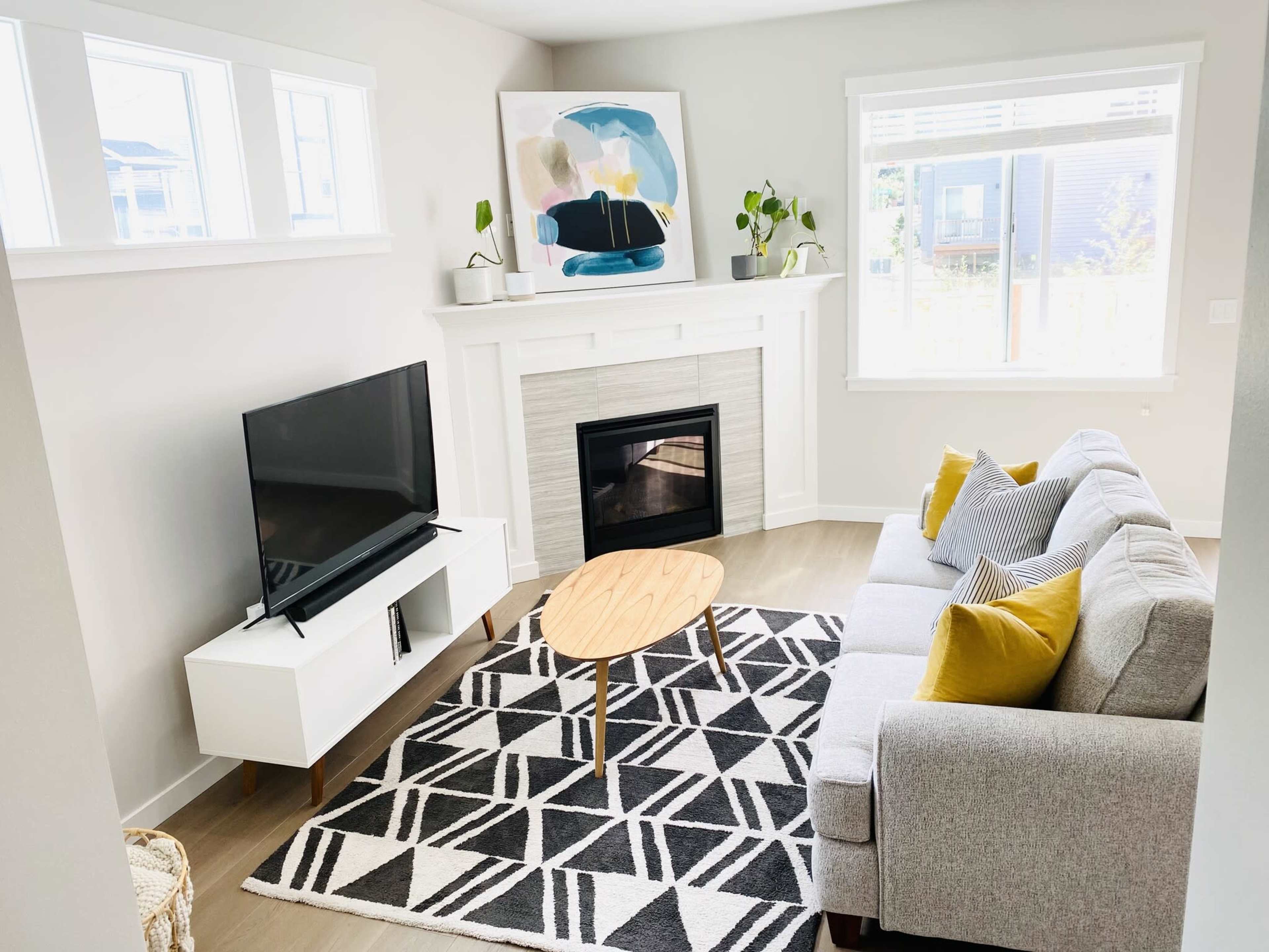A modern living room features a gray sofa, a wooden coffee table, a television on a white media console, and a fireplace beneath a large artwork, with natural light coming through the windows.