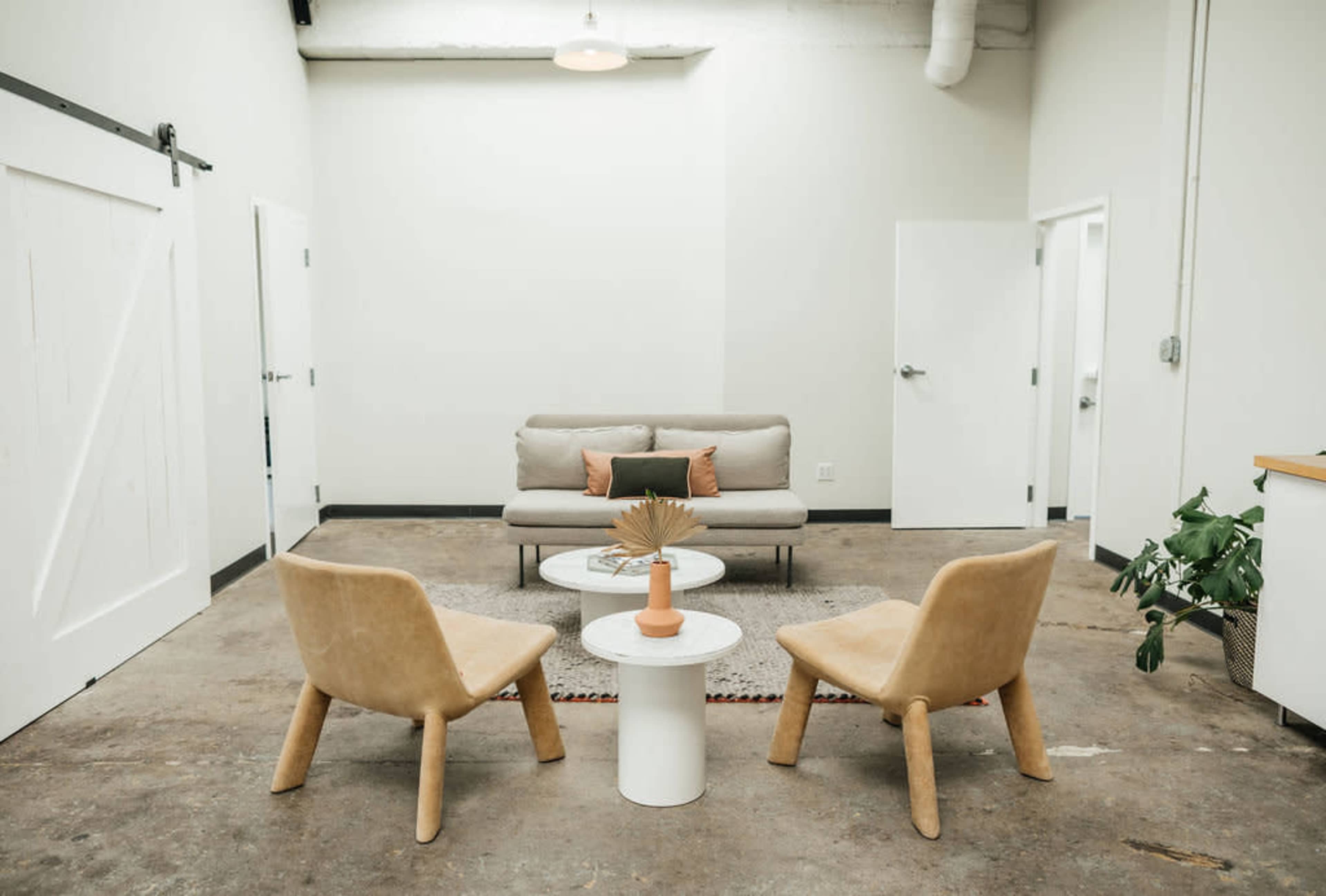 A minimalist lounge area features a light gray sofa, two tan chairs, a round coffee table with a decorative item, and neutral-colored walls.