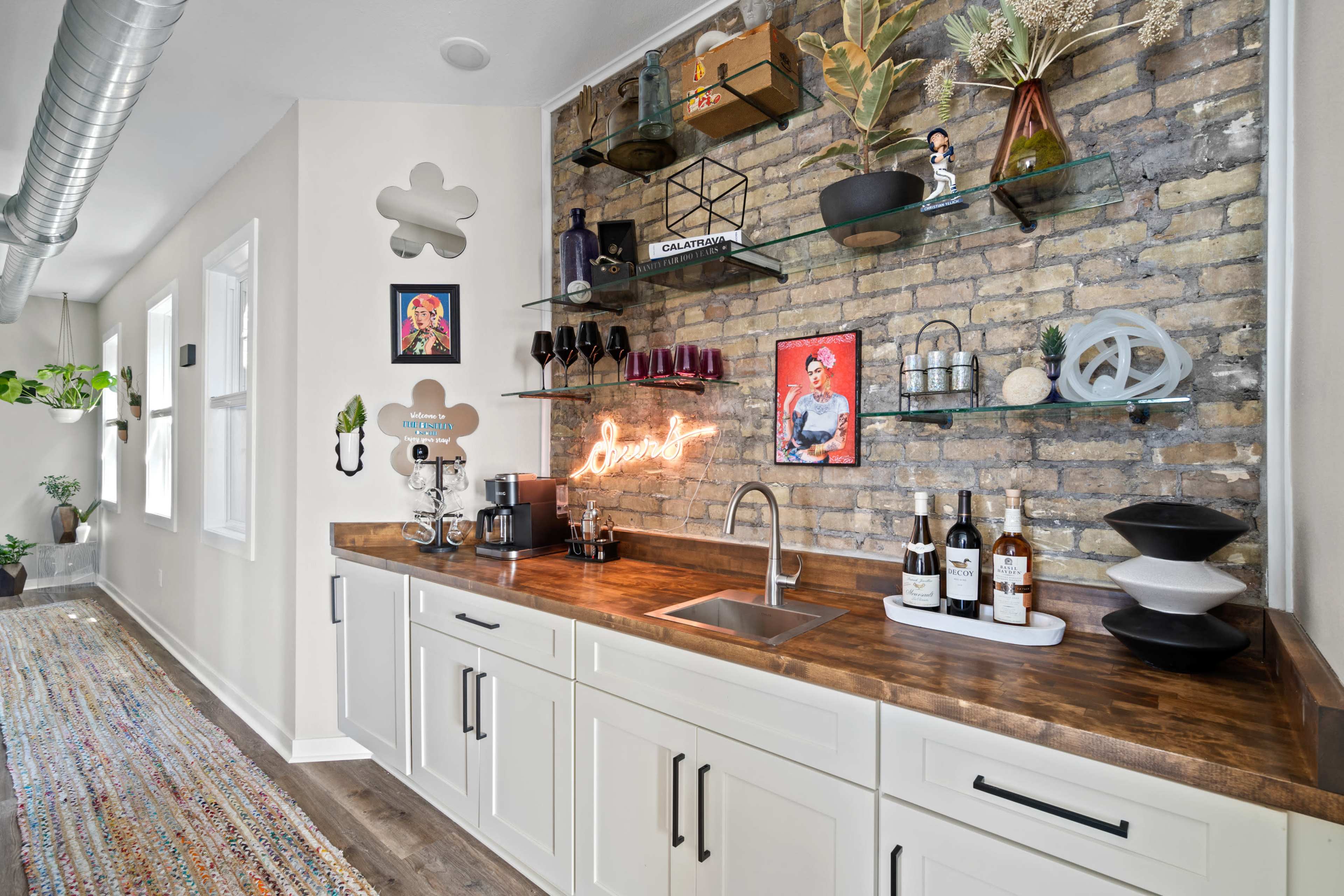 The image shows a modern kitchen bar area featuring a wooden countertop, open shelving with decorative items, a sink, and various bottles of wine displayed.