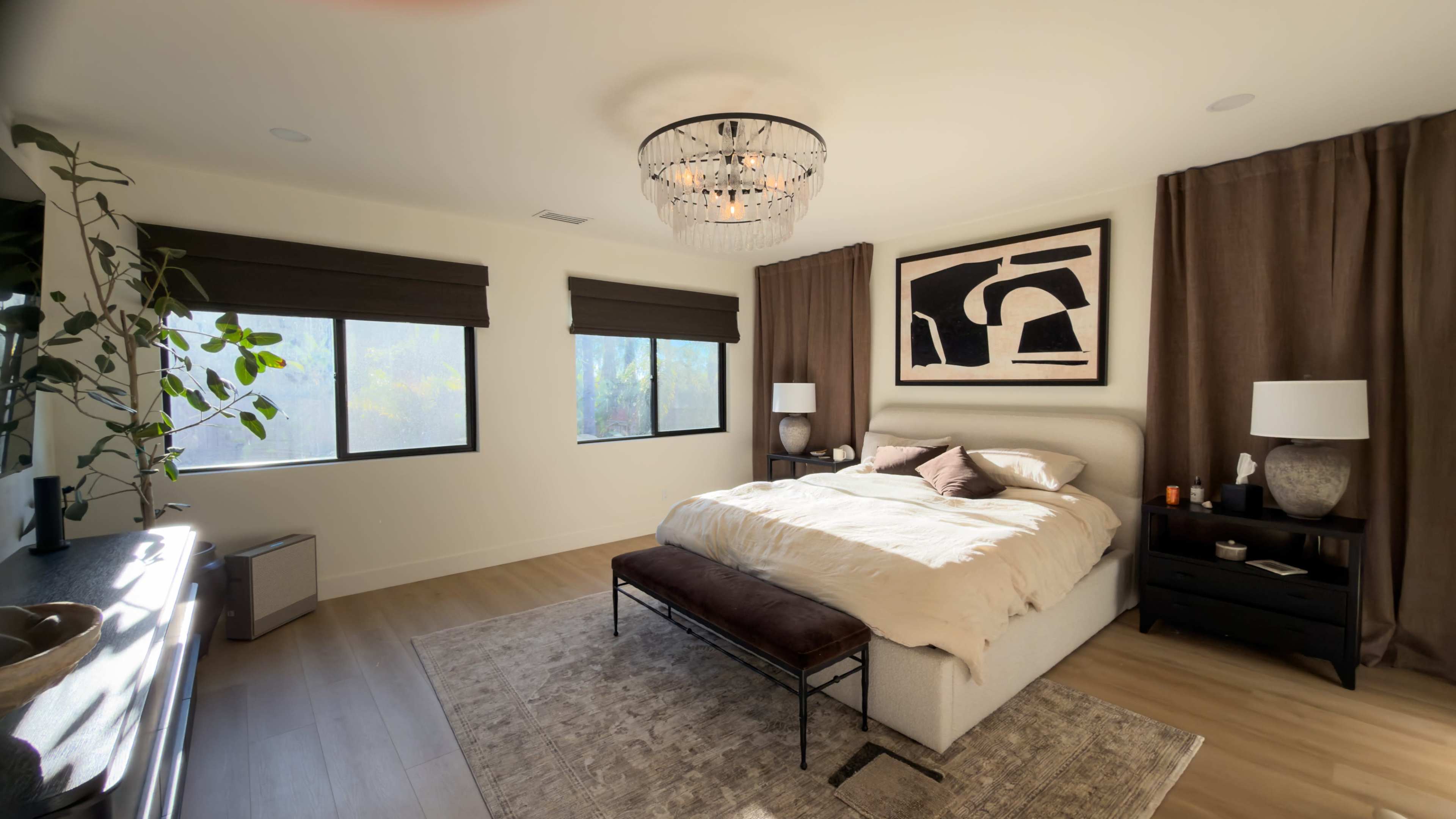 The bedroom features a large bed with a neutral-colored comforter, two bedside lamps, large windows with brown shades, and a piece of abstract art on the wall.