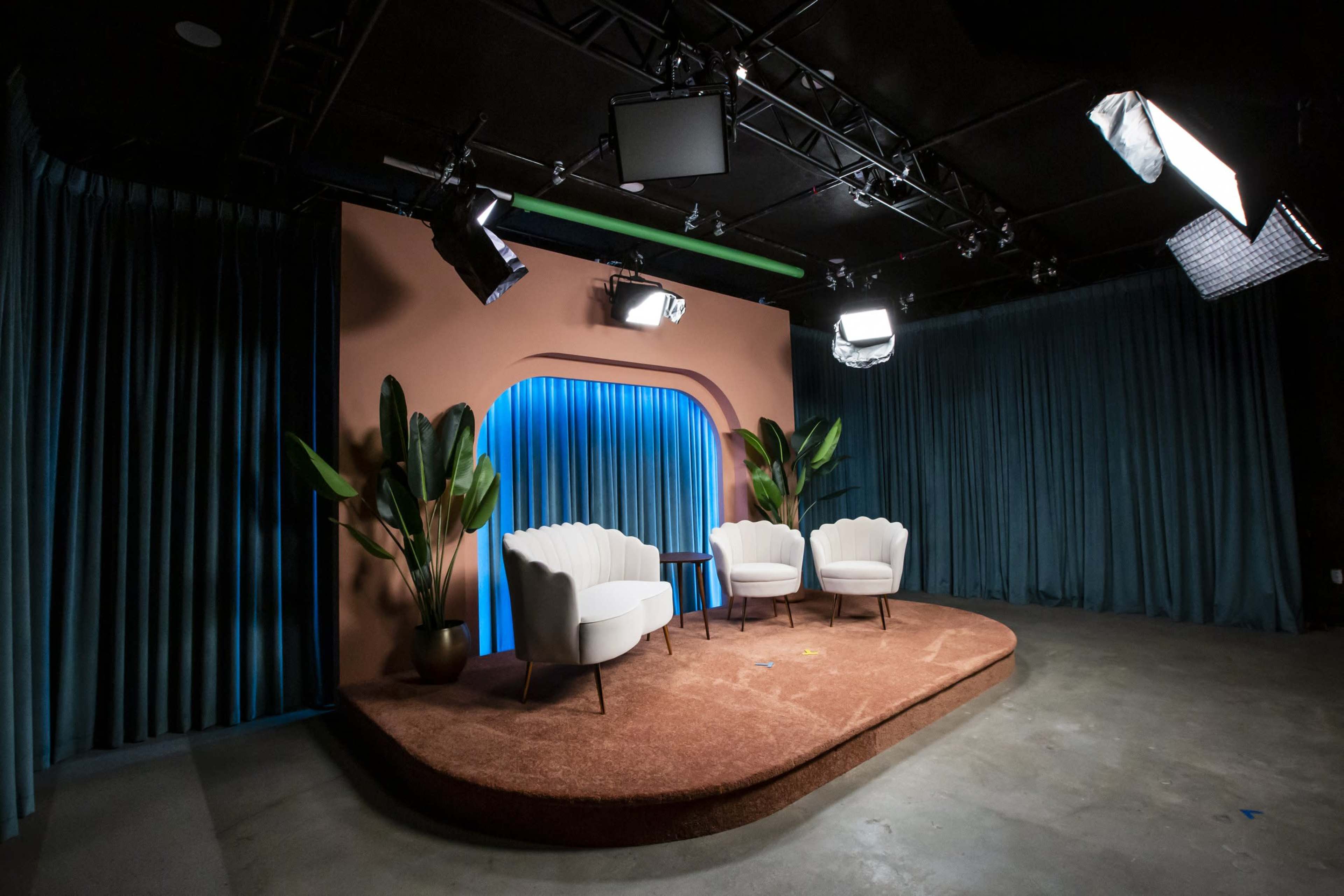 The image shows a modern set with three white chairs on a brown carpet, flanked by tall green plants and illuminated by multiple studio lights against a backdrop of blue and pink curtains.