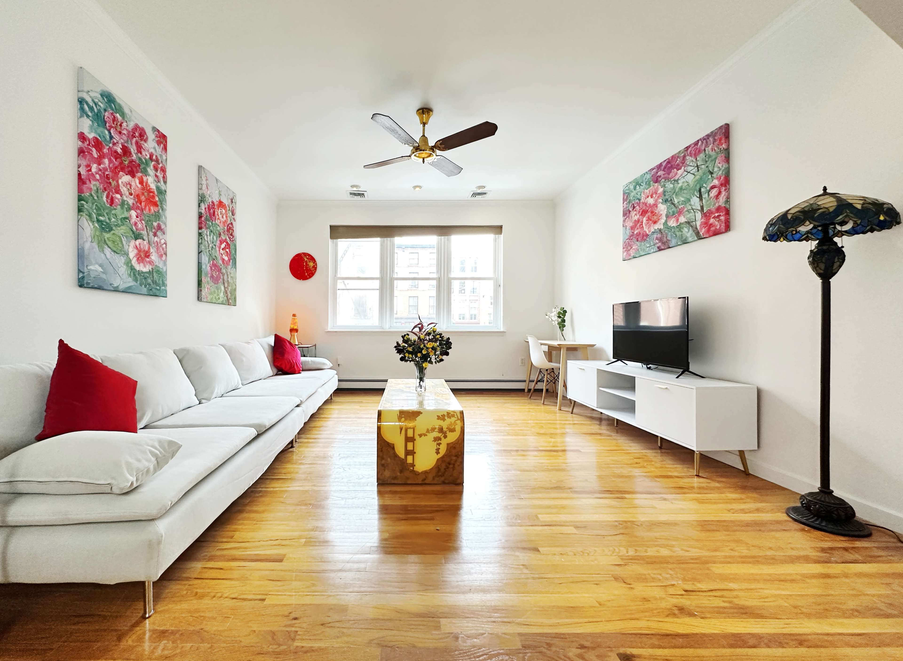 A bright living room featuring a white sofa, a wooden table with a lamp, and colorful floral artwork on the walls.