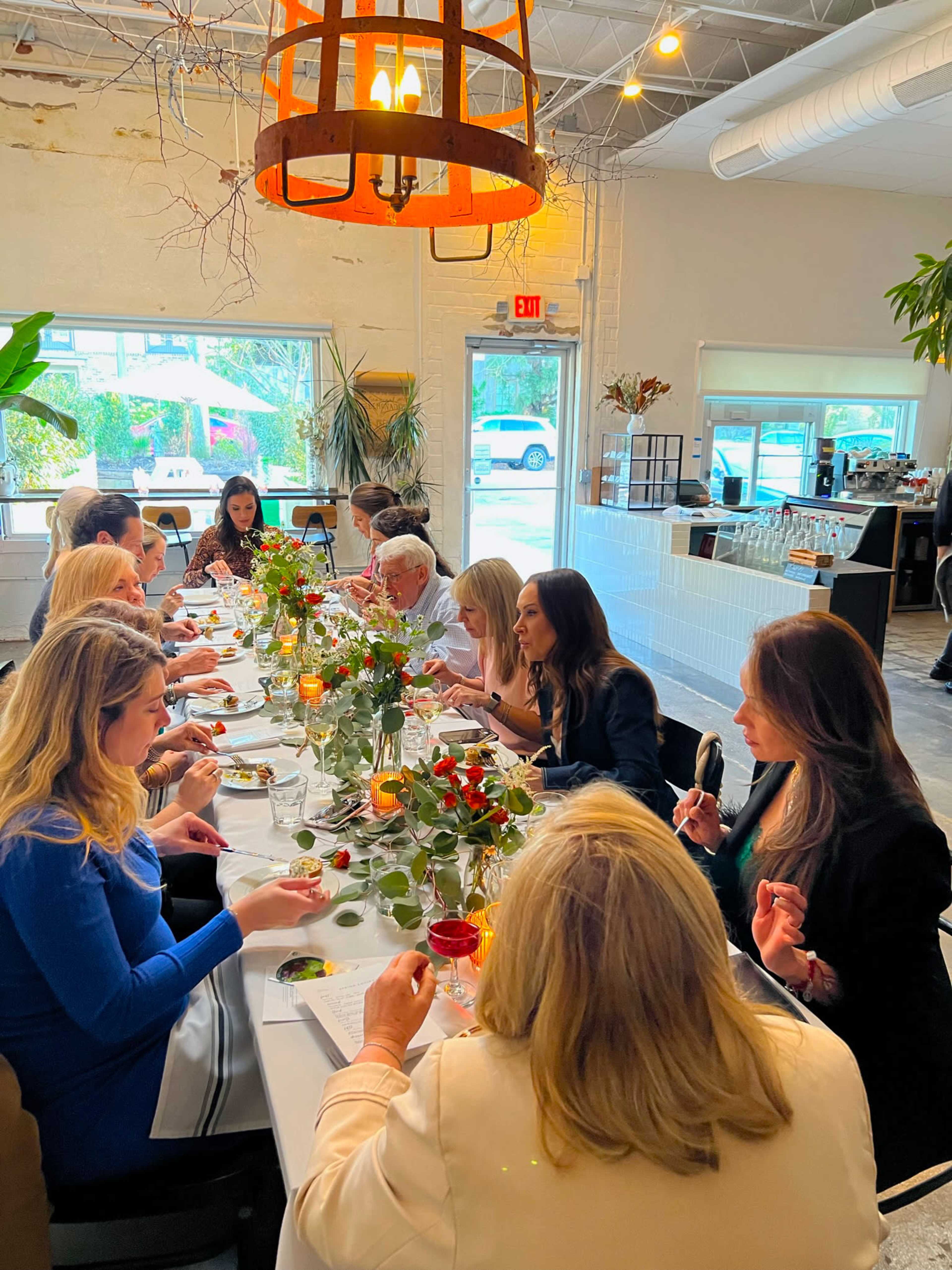 A group of people is seated at a long table adorned with flowers, enjoying a meal in a brightly lit restaurant.