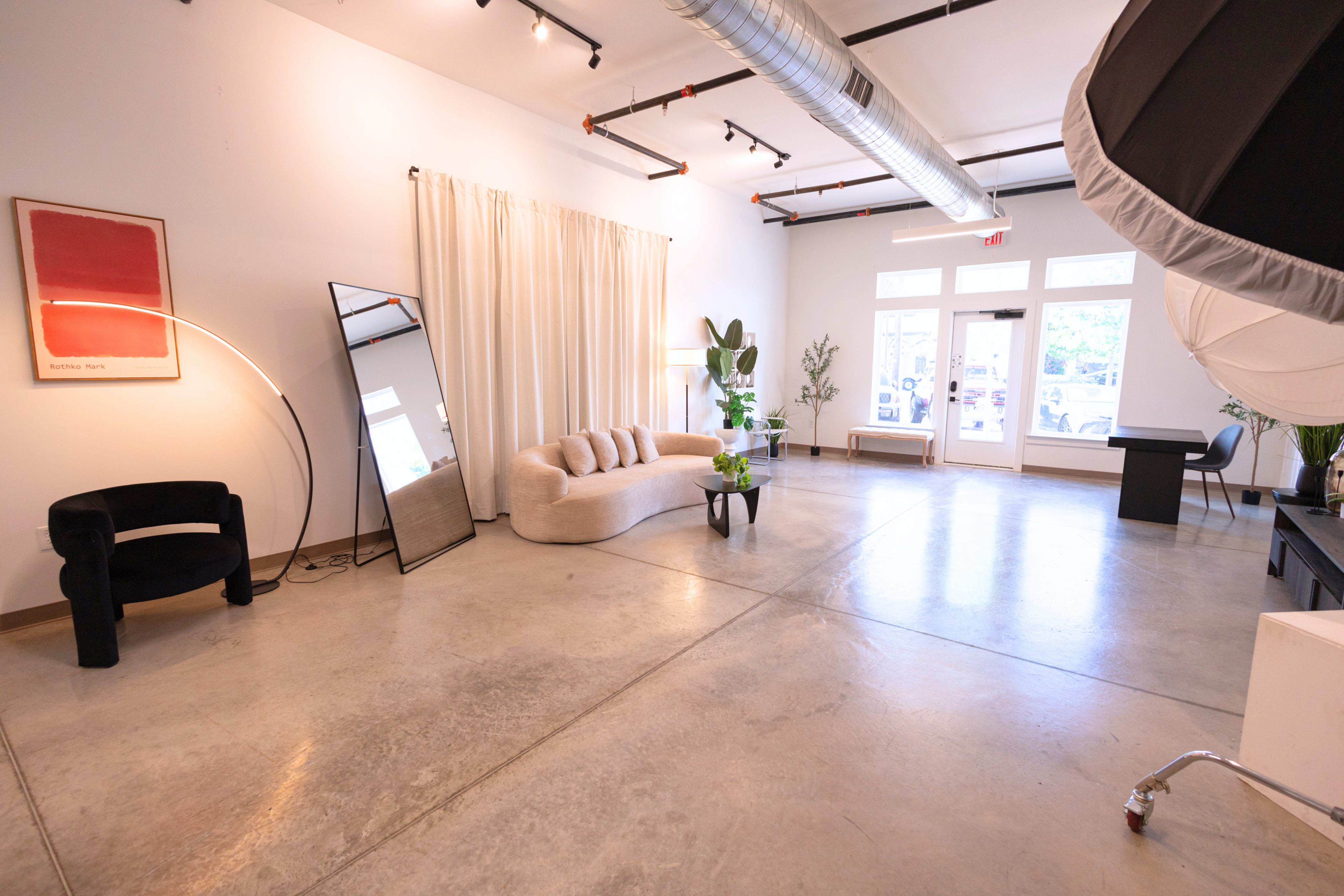 The image shows a spacious and modern interior featuring a light-colored couch, a large mirror, a potted plant, and natural light coming through large windows.