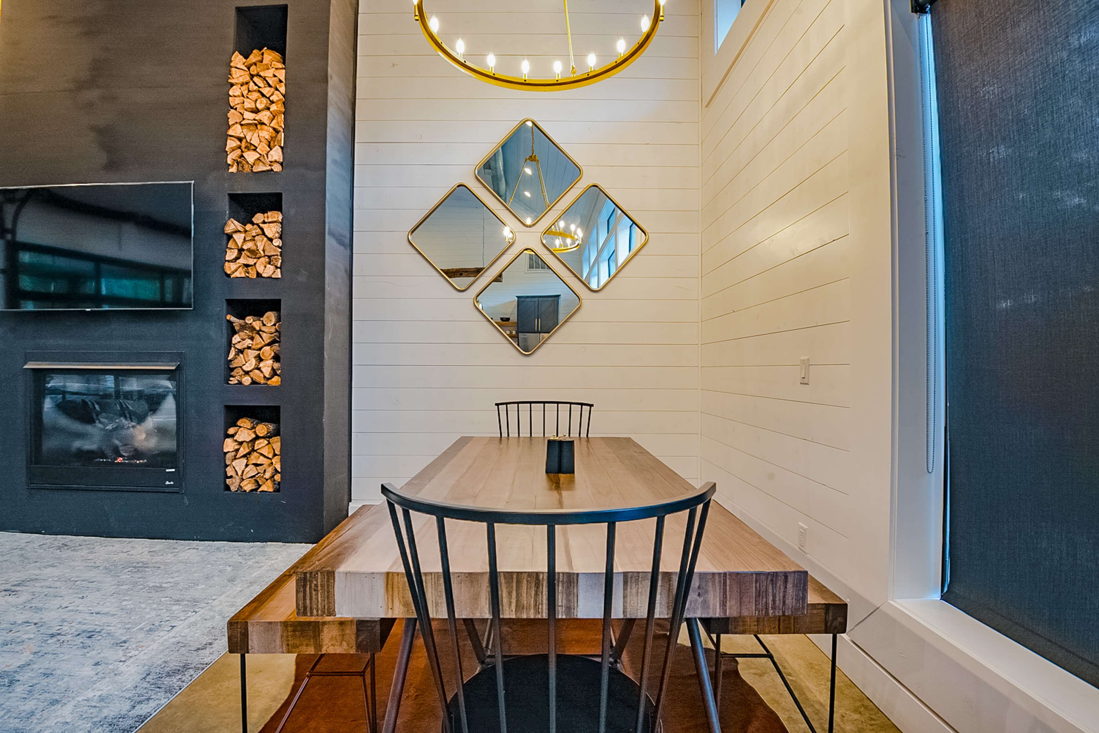 A modern dining area featuring a wooden table with black chairs, a large mirror, and a stacked wood wall near a fireplace.