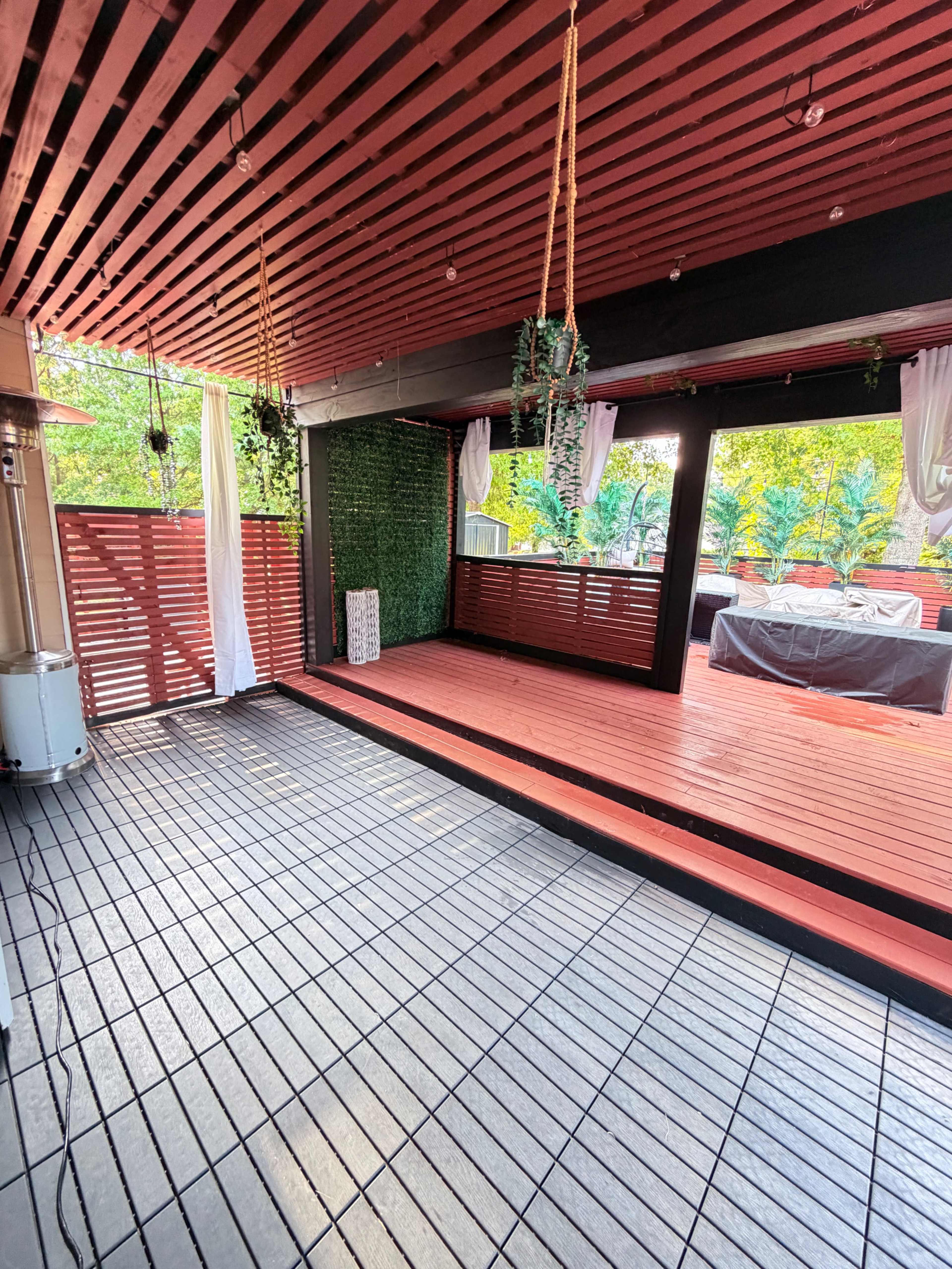 The image shows a wooden deck area with red-painted beams and a grid-patterned floor, surrounded by greenery and seating arrangements.