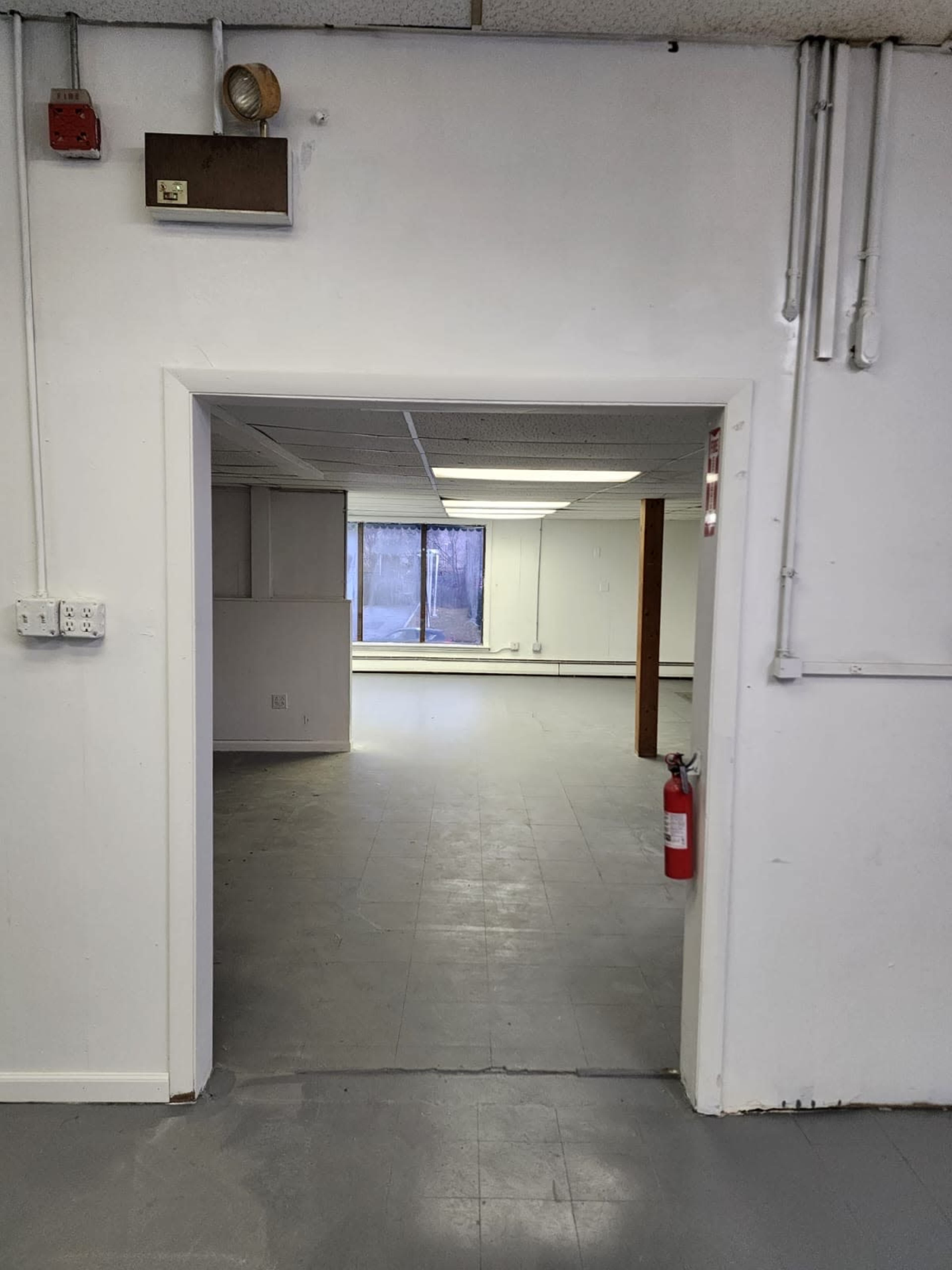 The image shows an empty room with a doorway leading into another large, similarly vacant space illuminated by natural light from a window.