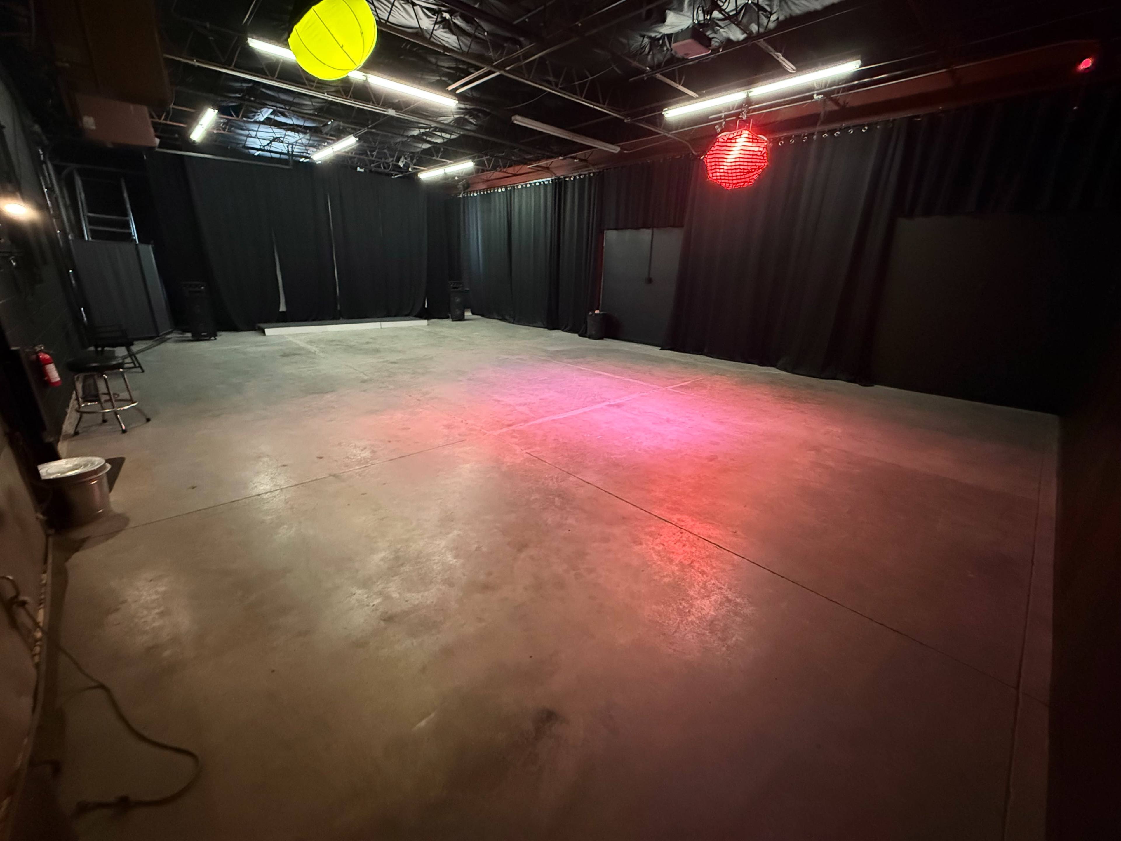 The image shows an empty performance space with a concrete floor and black curtains surrounding the area, illuminated by colored lights.