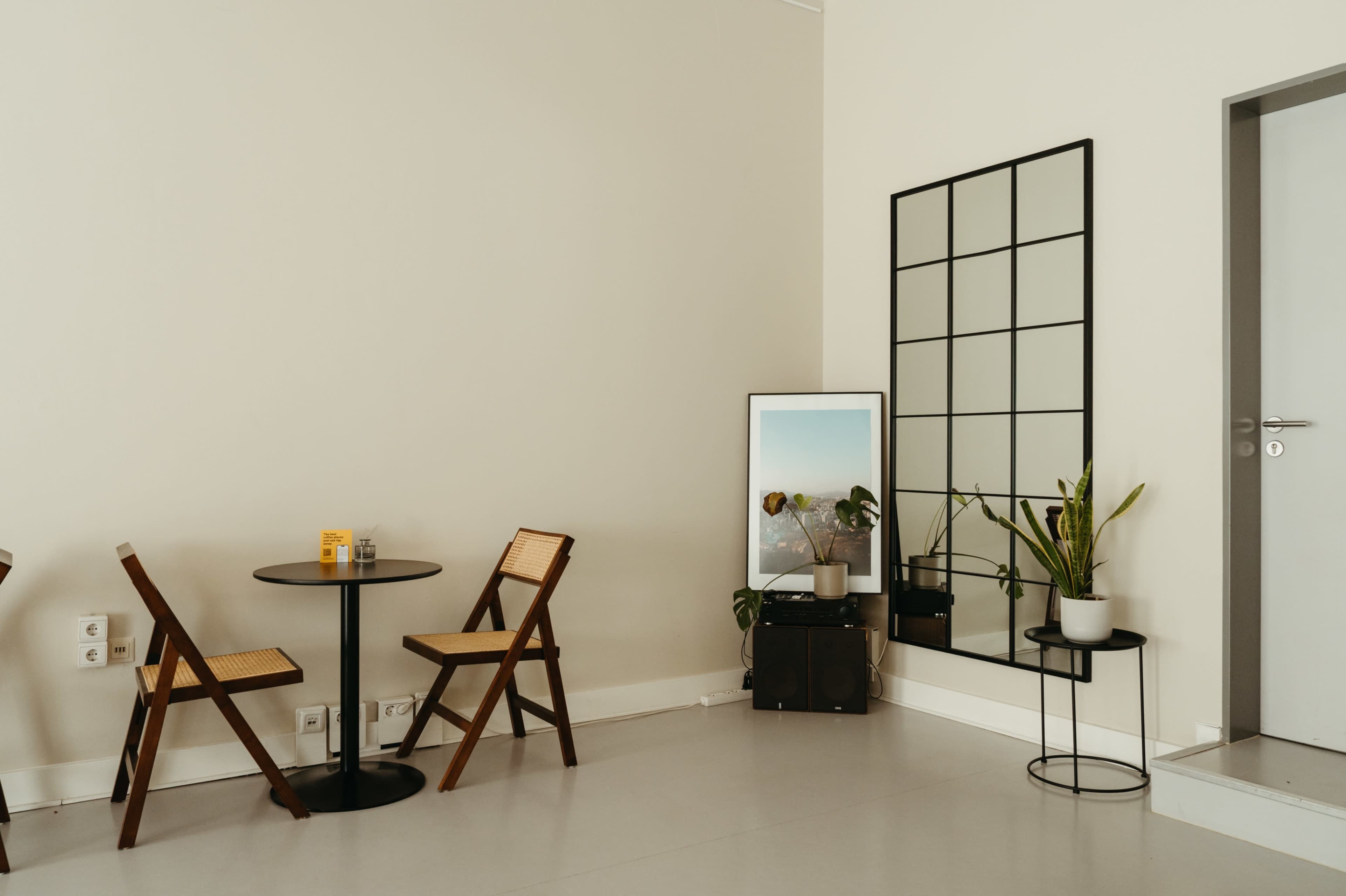 The image shows a minimalist interior space with two wooden folding chairs and a round table next to a wall featuring a large mirror and a potted plant.