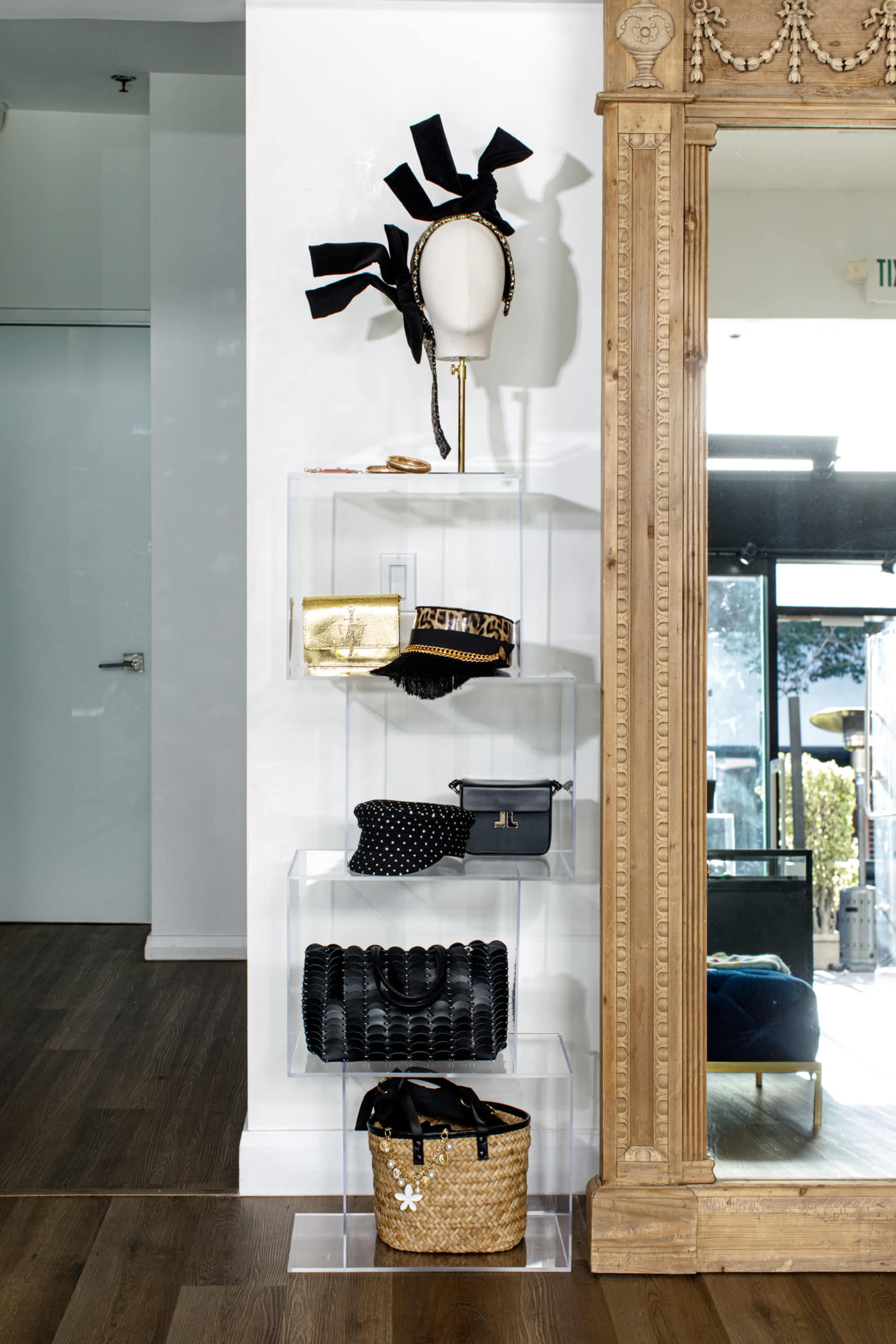 A clear display shelf showcases a mannequin head with a black accessory, various handbags, and a gold clutch against a backdrop featuring a large ornate mirror.