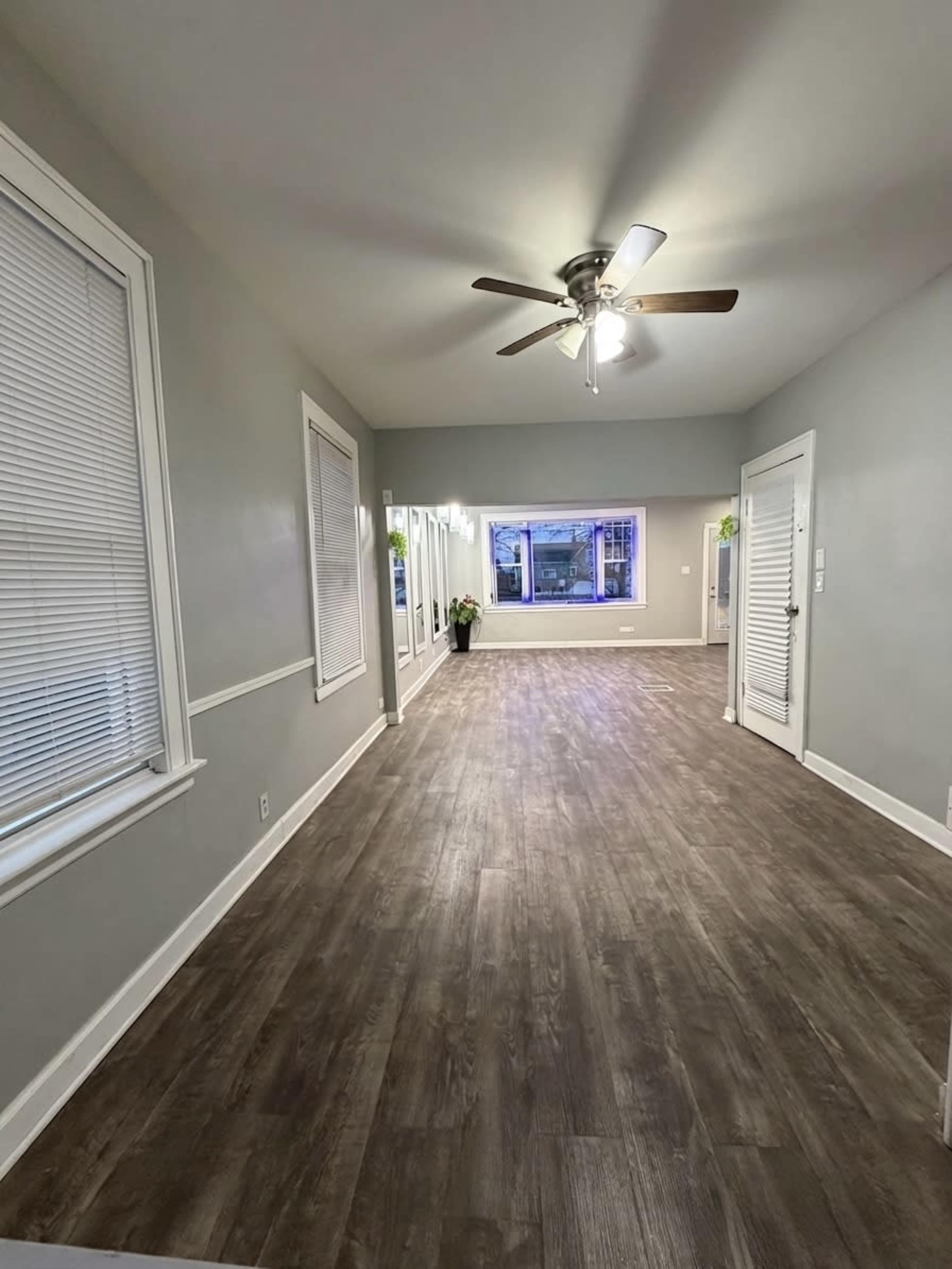 The image shows a spacious room with a ceiling fan, light gray walls, and a wooden floor, featuring large windows and a door on the right side.