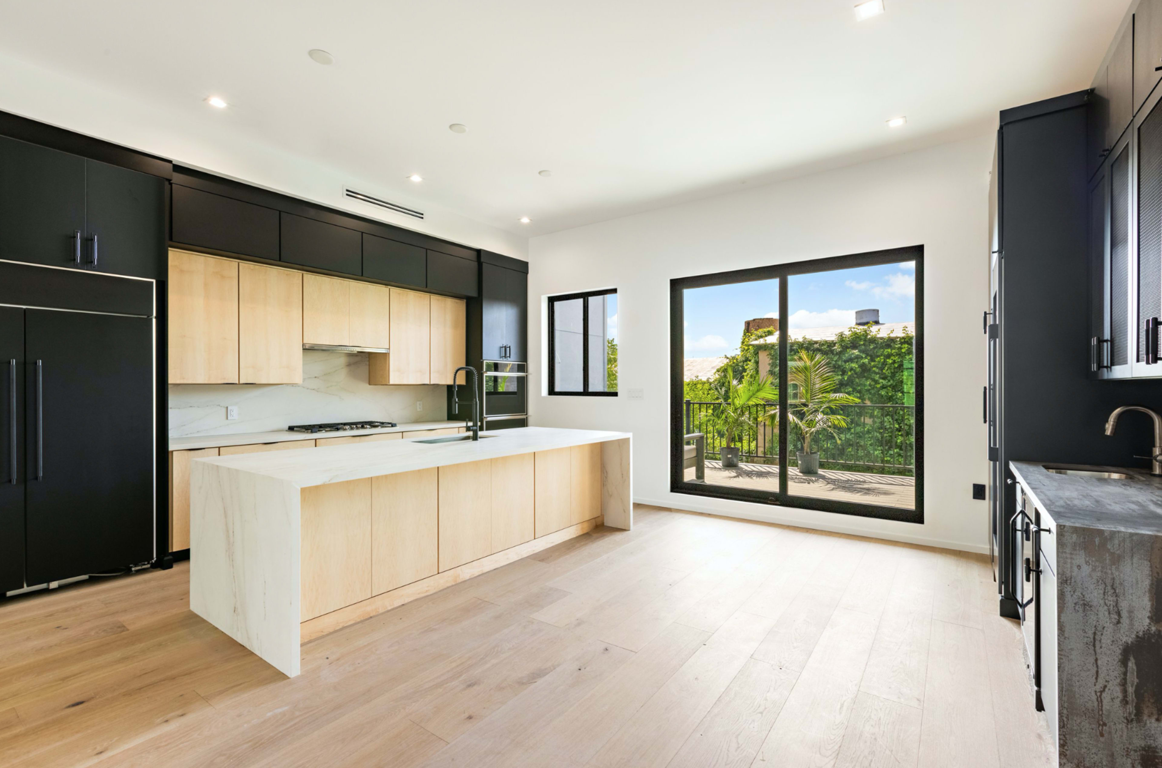 A modern kitchen features a large island with a light wood finish, sleek black cabinetry, and a large window overlooking a balcony with greenery.