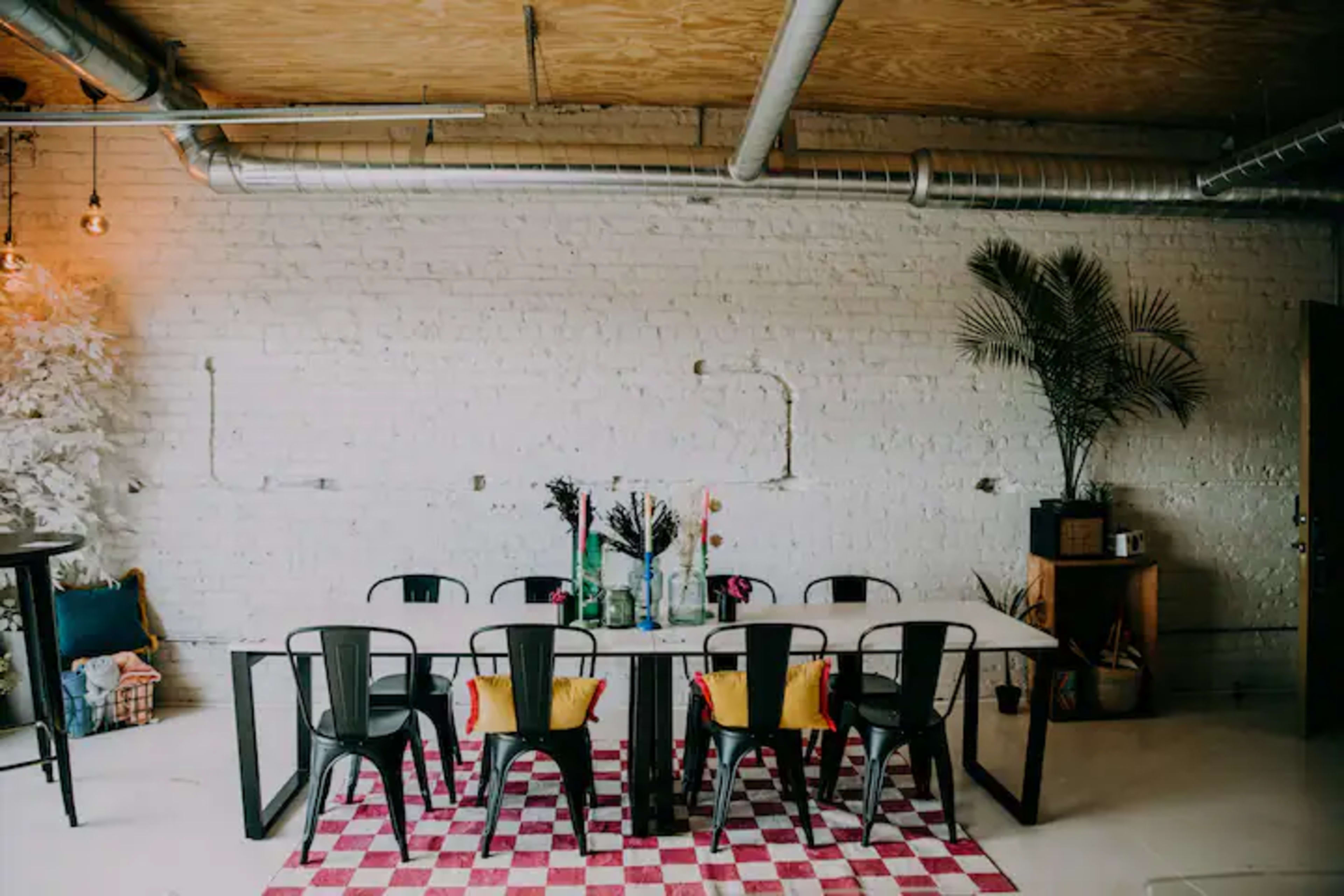 A long dining table surrounded by black chairs is set against a white brick wall, with colorful decorations and a patterned rug underneath.