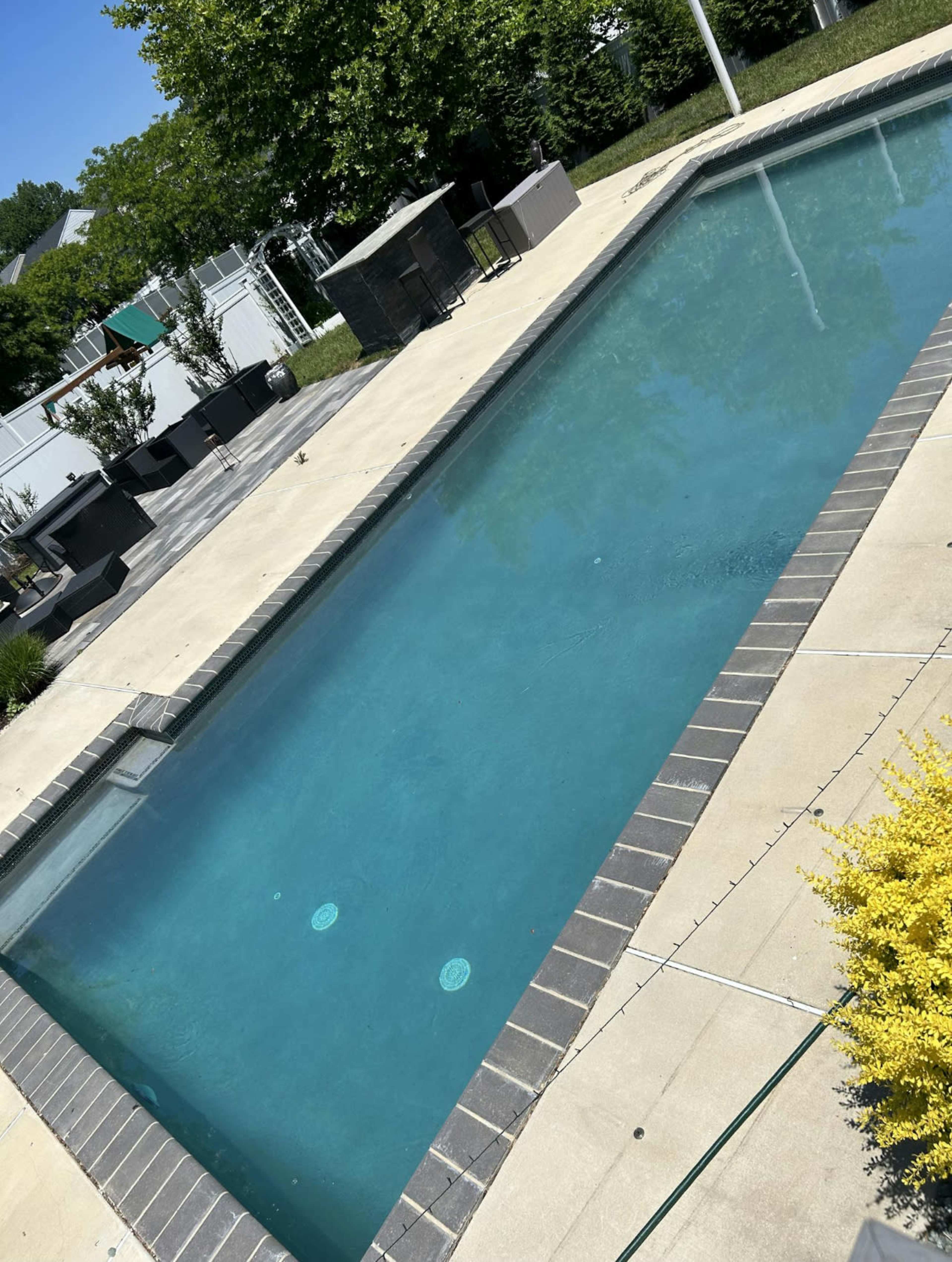 The image shows a rectangular swimming pool surrounded by a paved deck and outdoor furniture in a well-maintained backyard.