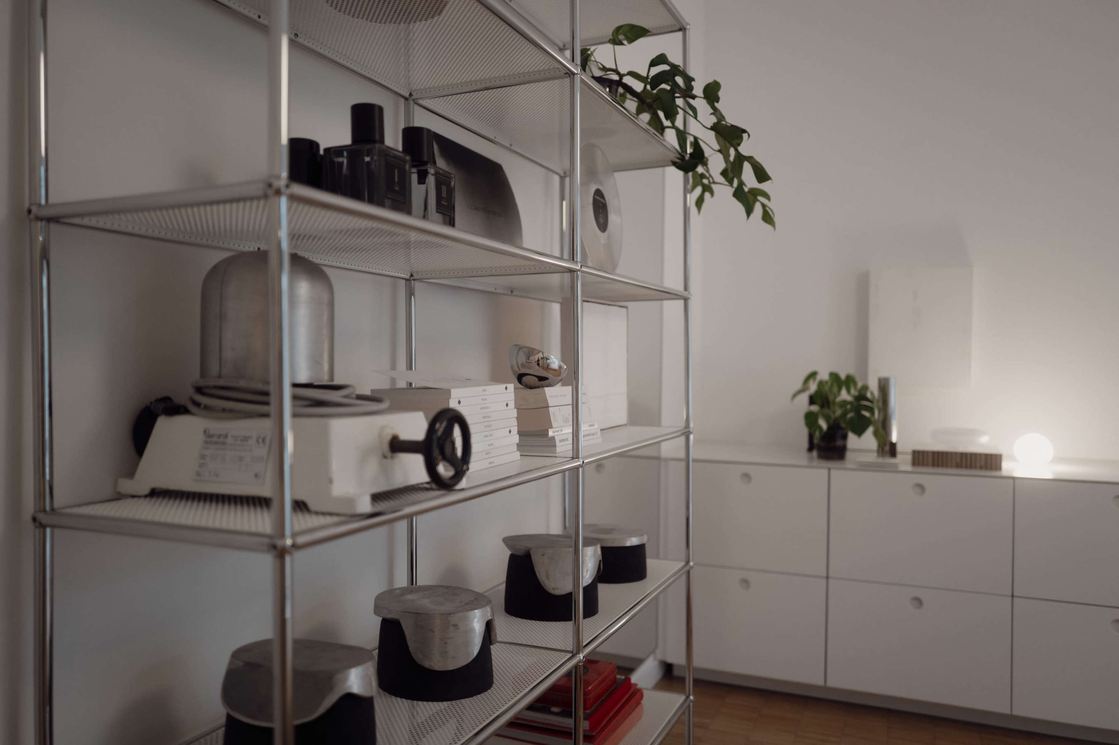 A metal shelving unit displays various kitchen tools, books, and a potted plant in a minimalist interior space.