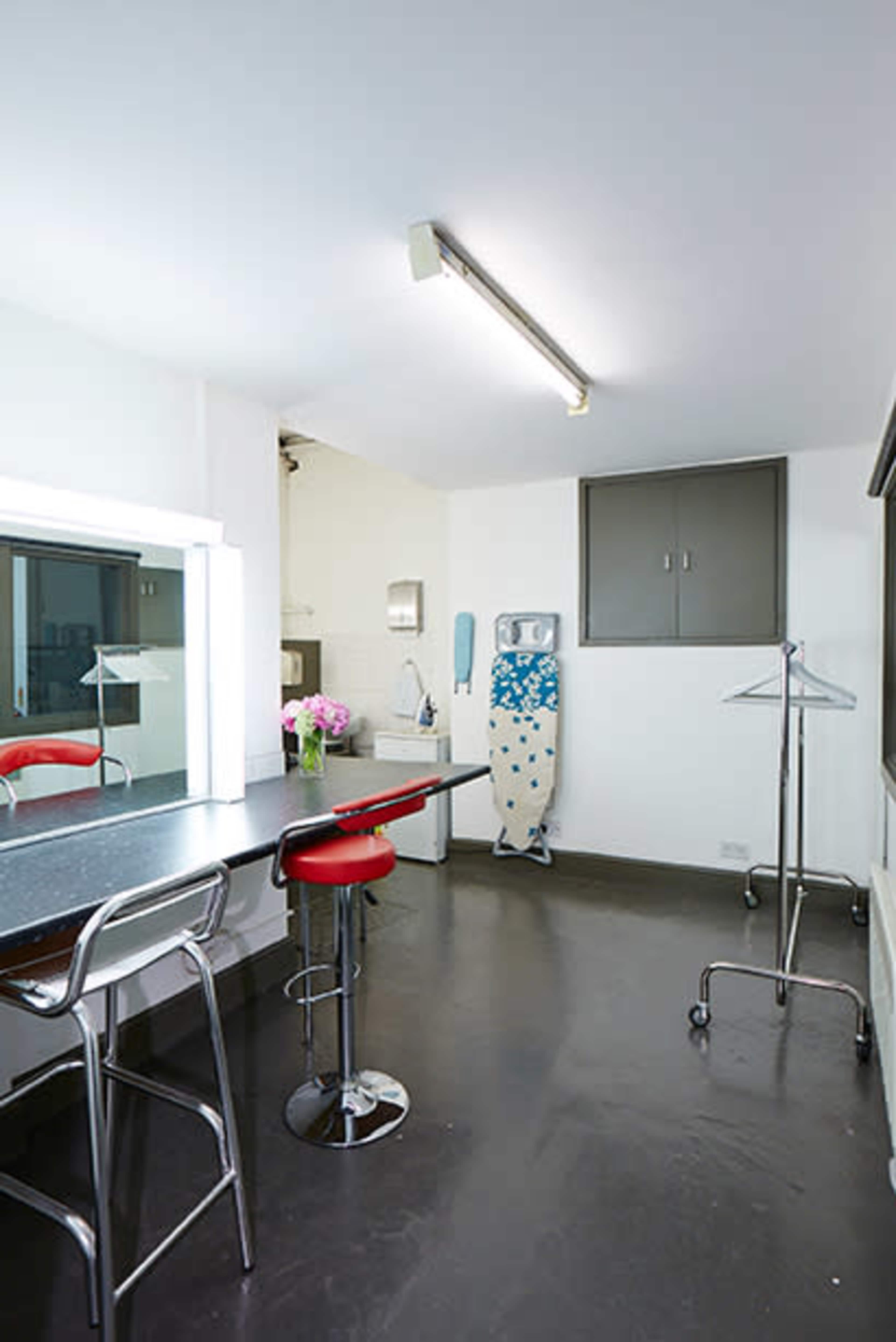A minimalist room with a black table, red bar stools, an ironing board, a clothing rack, and a window displaying a view of a kitchen area.
