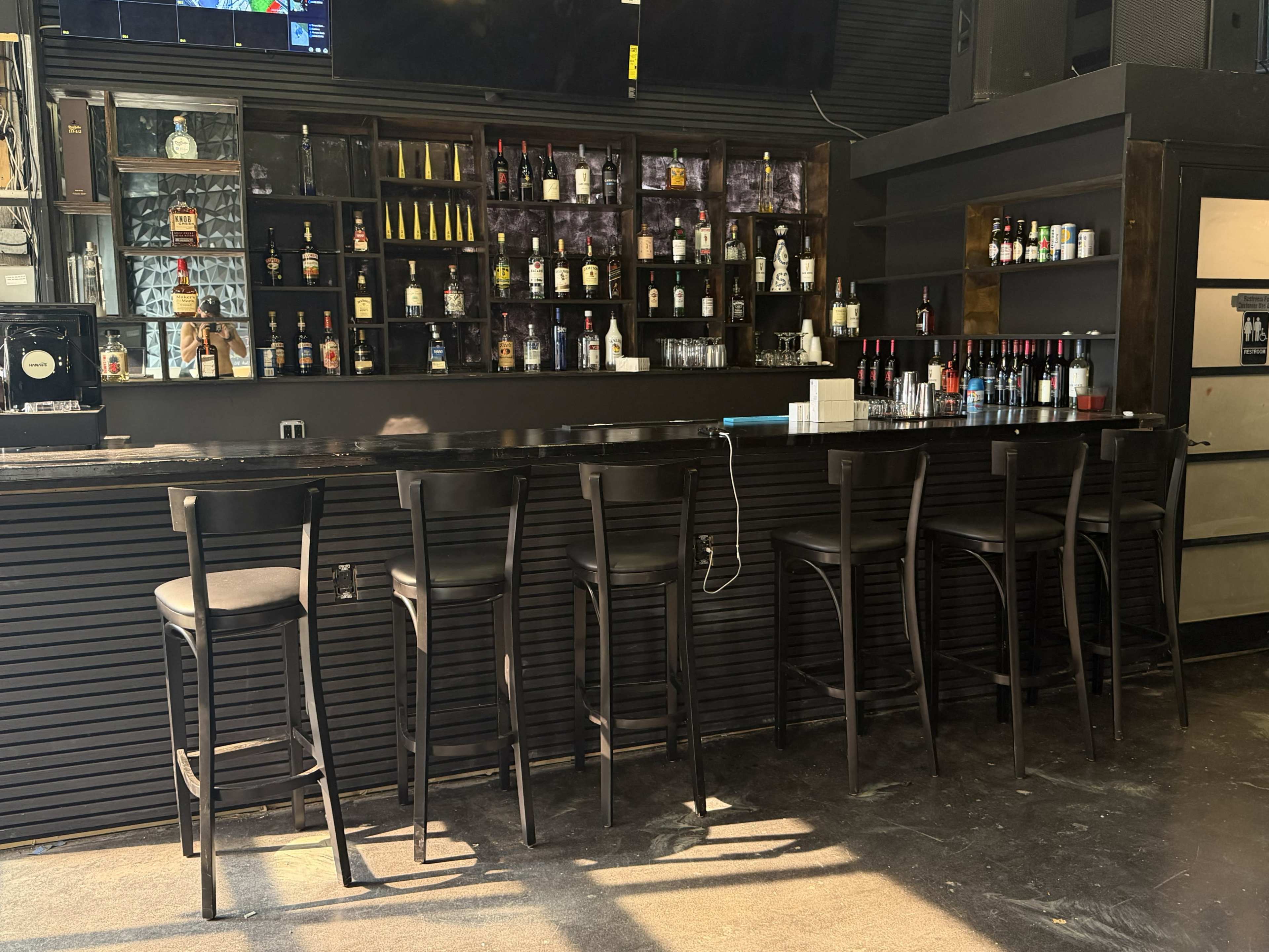 The image shows an empty bar area with high stools arranged neatly in front of a dark wooden counter, against a backdrop of shelves filled with various bottles.