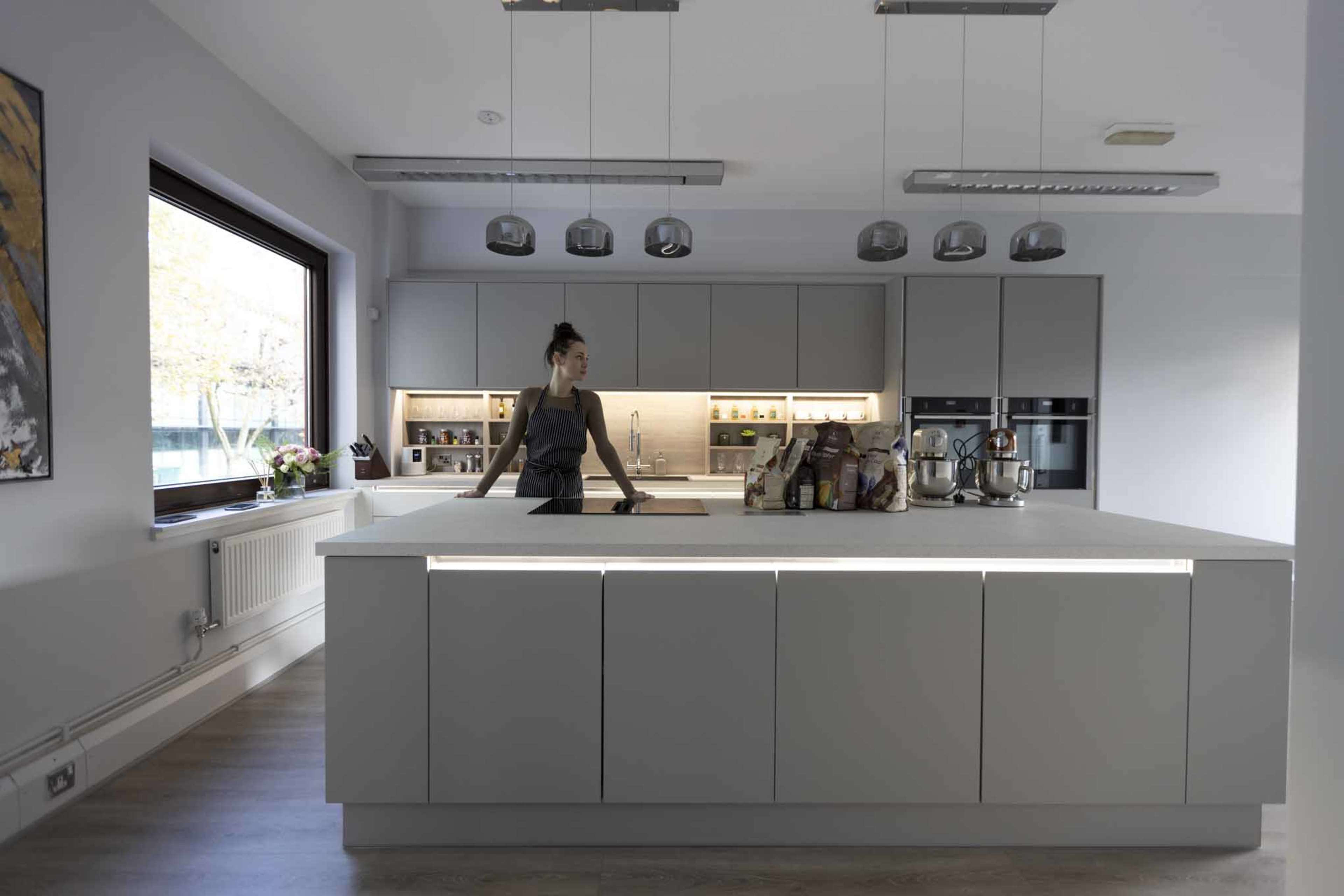 A woman stands at a modern kitchen island with a light-colored countertop, surrounded by sleek cabinetry and multiple pendant lights overhead.