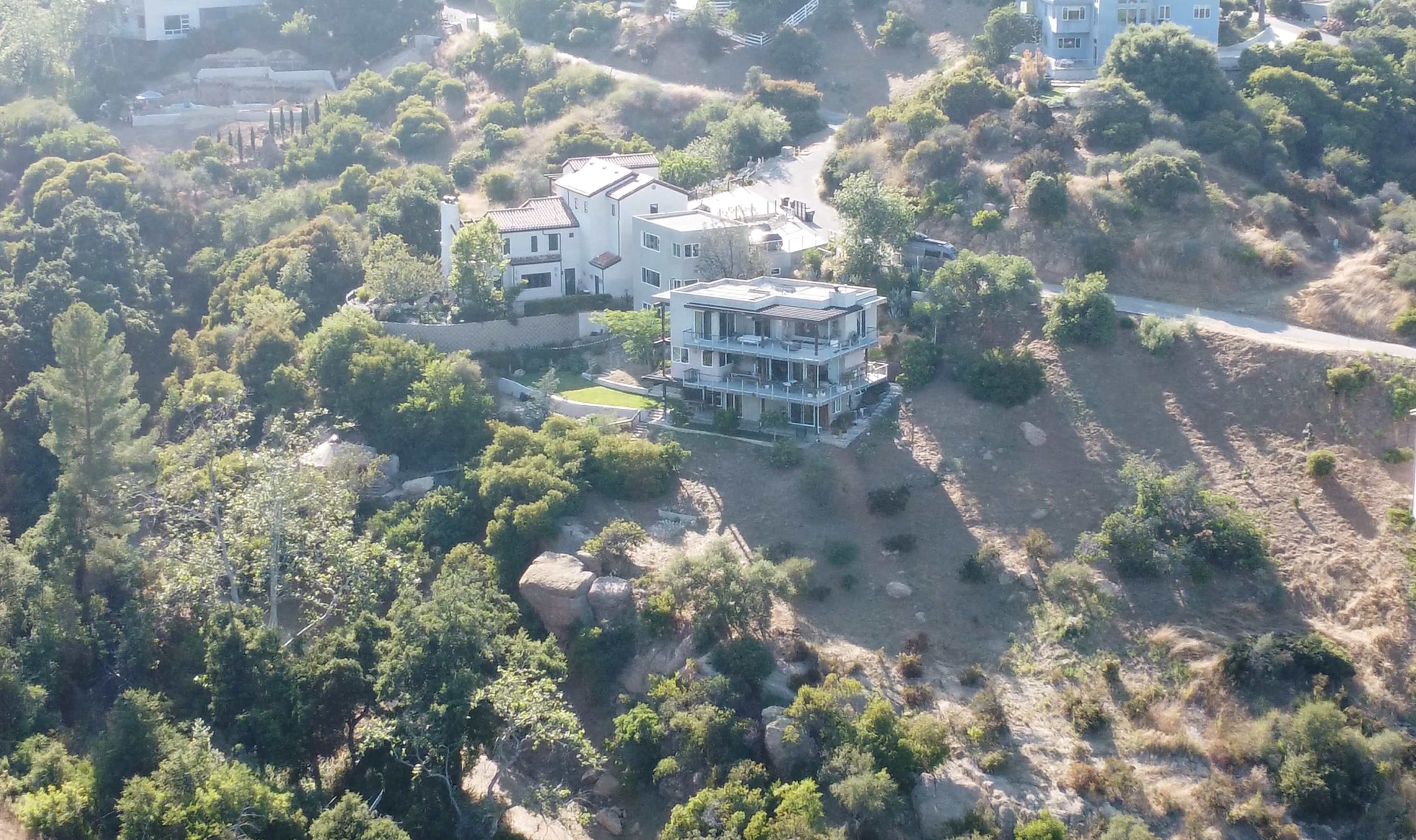A modern house sits on a hillside surrounded by greenery, with another building nearby.