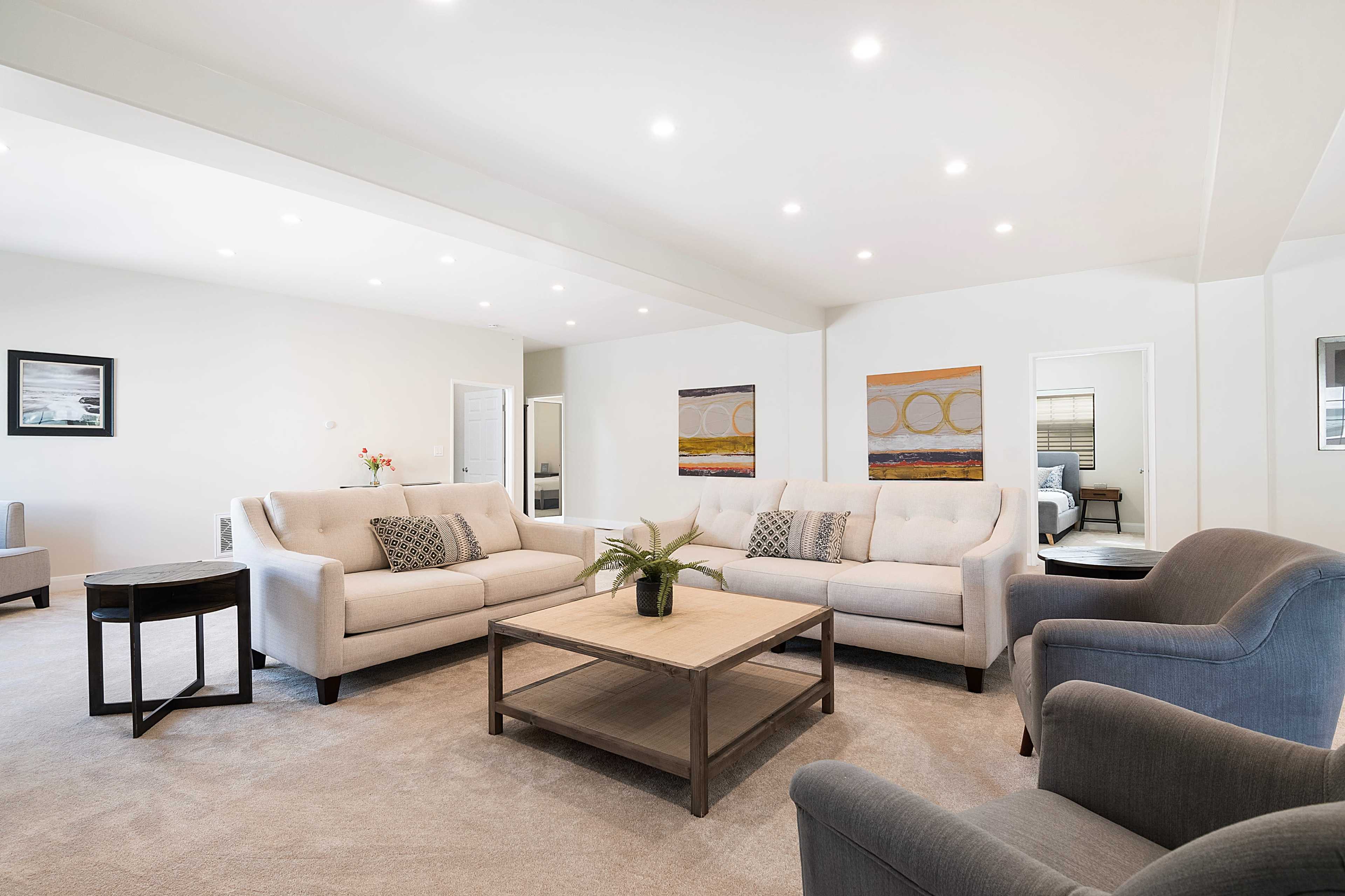 A spacious living room features light-colored sofas arranged around a wooden coffee table, with artwork on the walls and natural light illuminating the space.