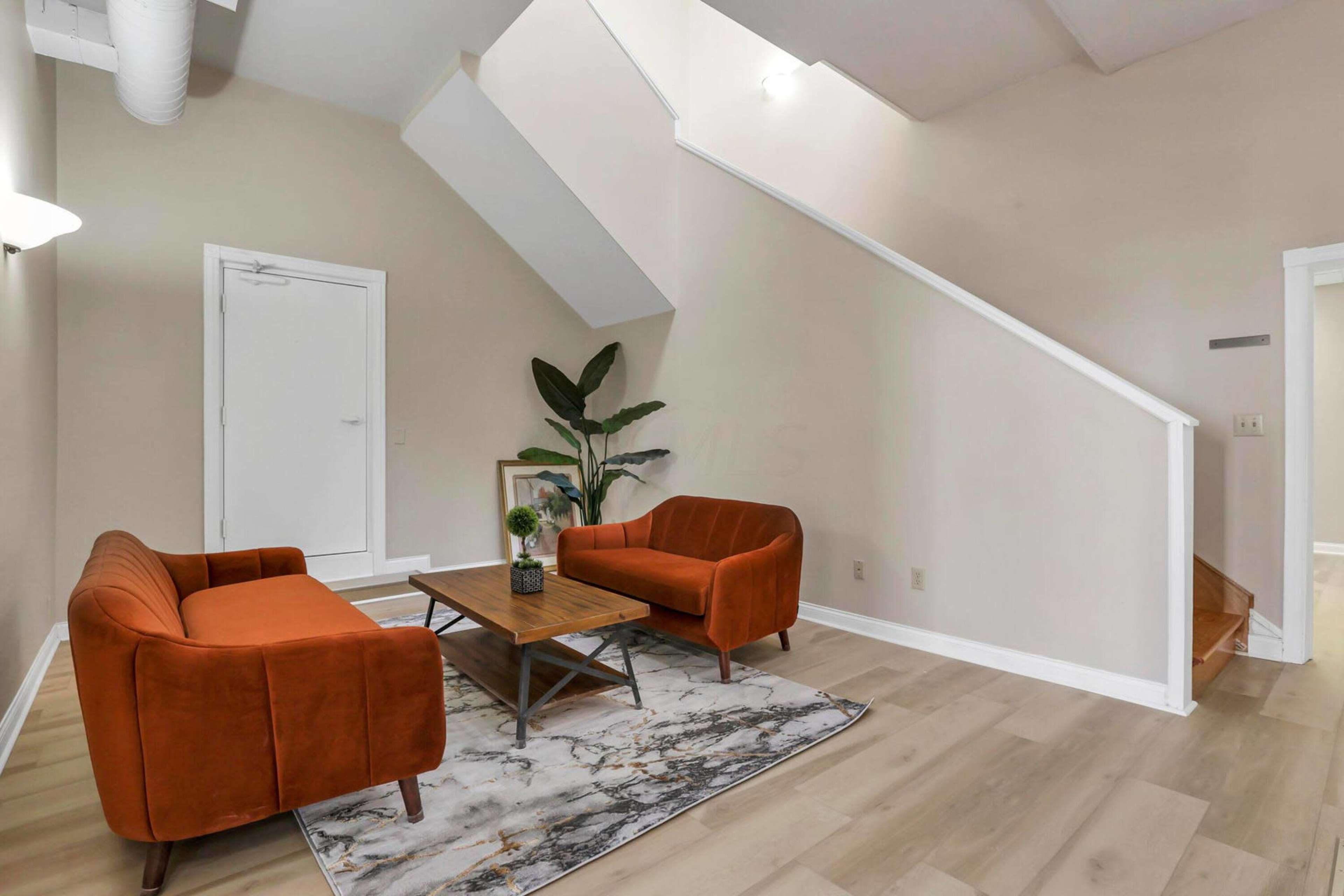 A living area with two orange sofas facing a wooden coffee table on a marble-patterned rug, adjacent to a staircase and a potted plant.