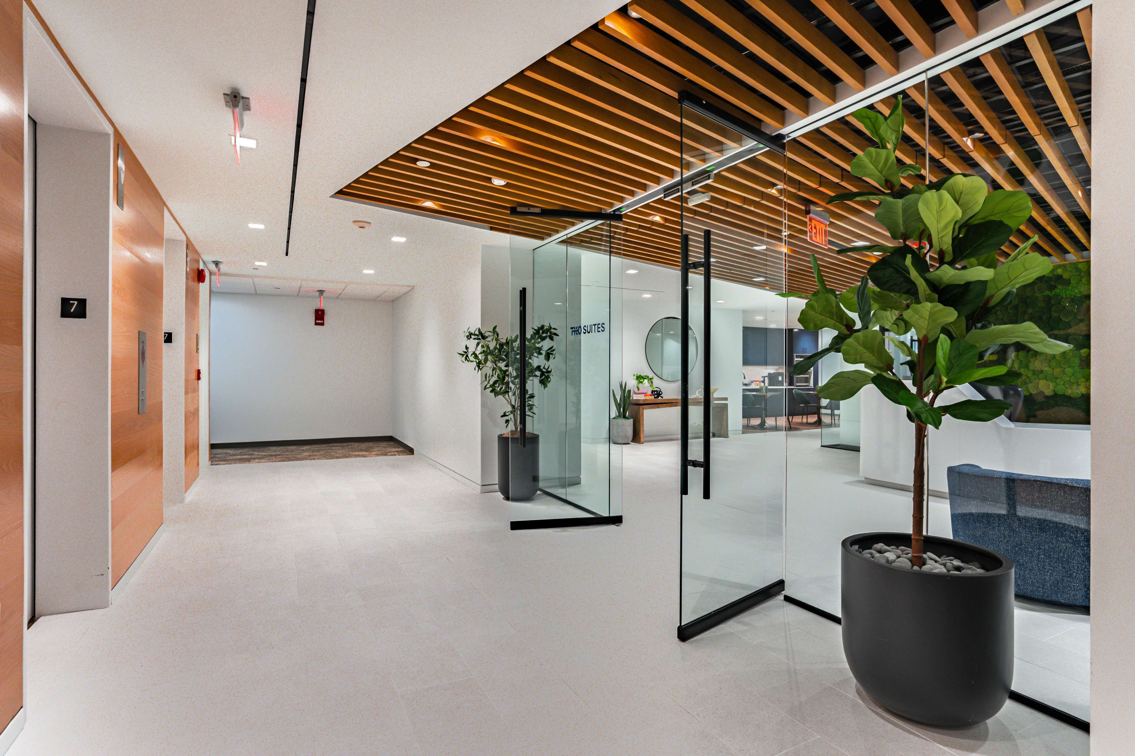 The image shows a modern hallway featuring an open glass door leading to a lounge area, with elevators on the left and potted plants along the walls.