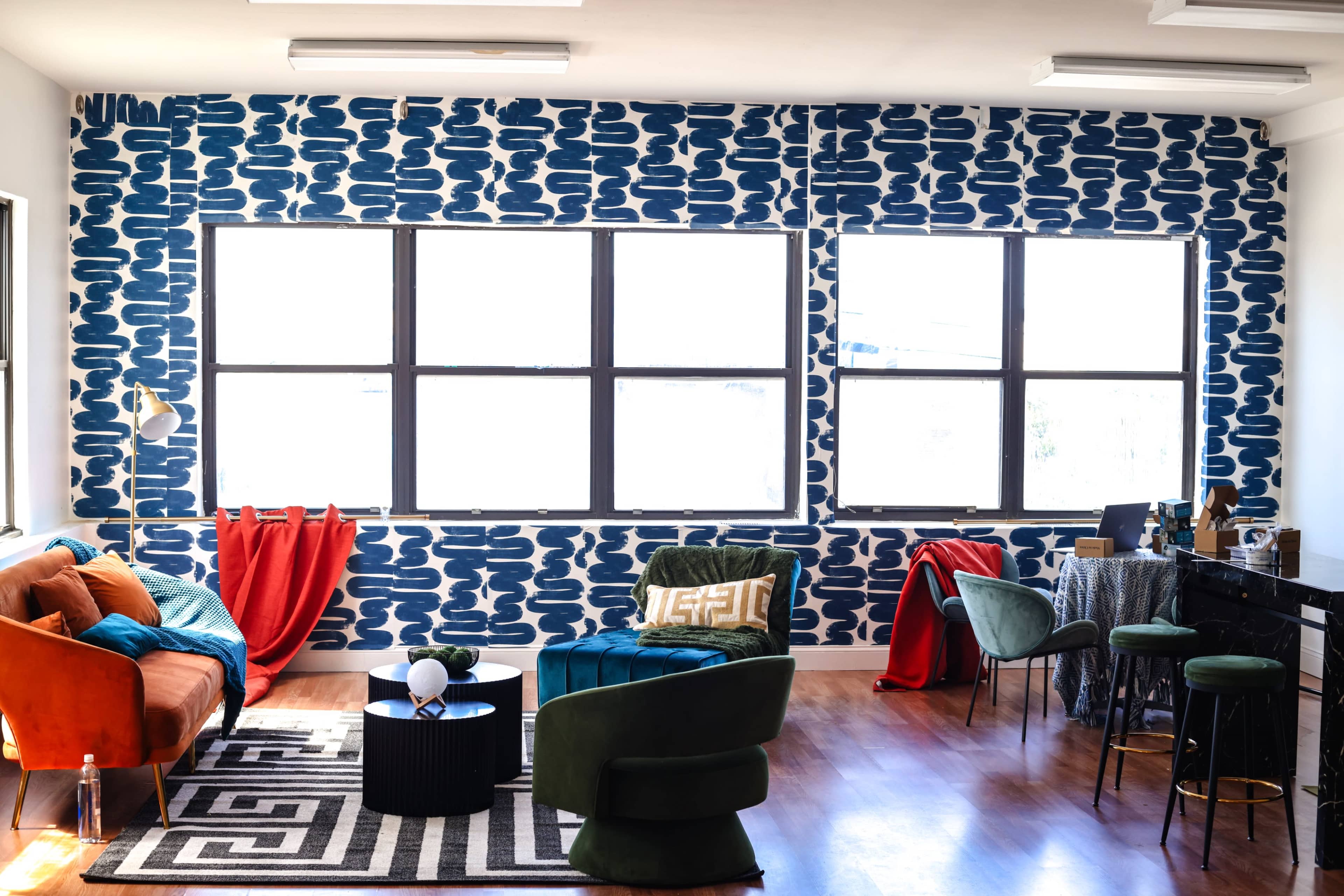 A spacious room features large windows with patterned blue wallpaper, colorful furniture, and a sleek dark countertop workspace.