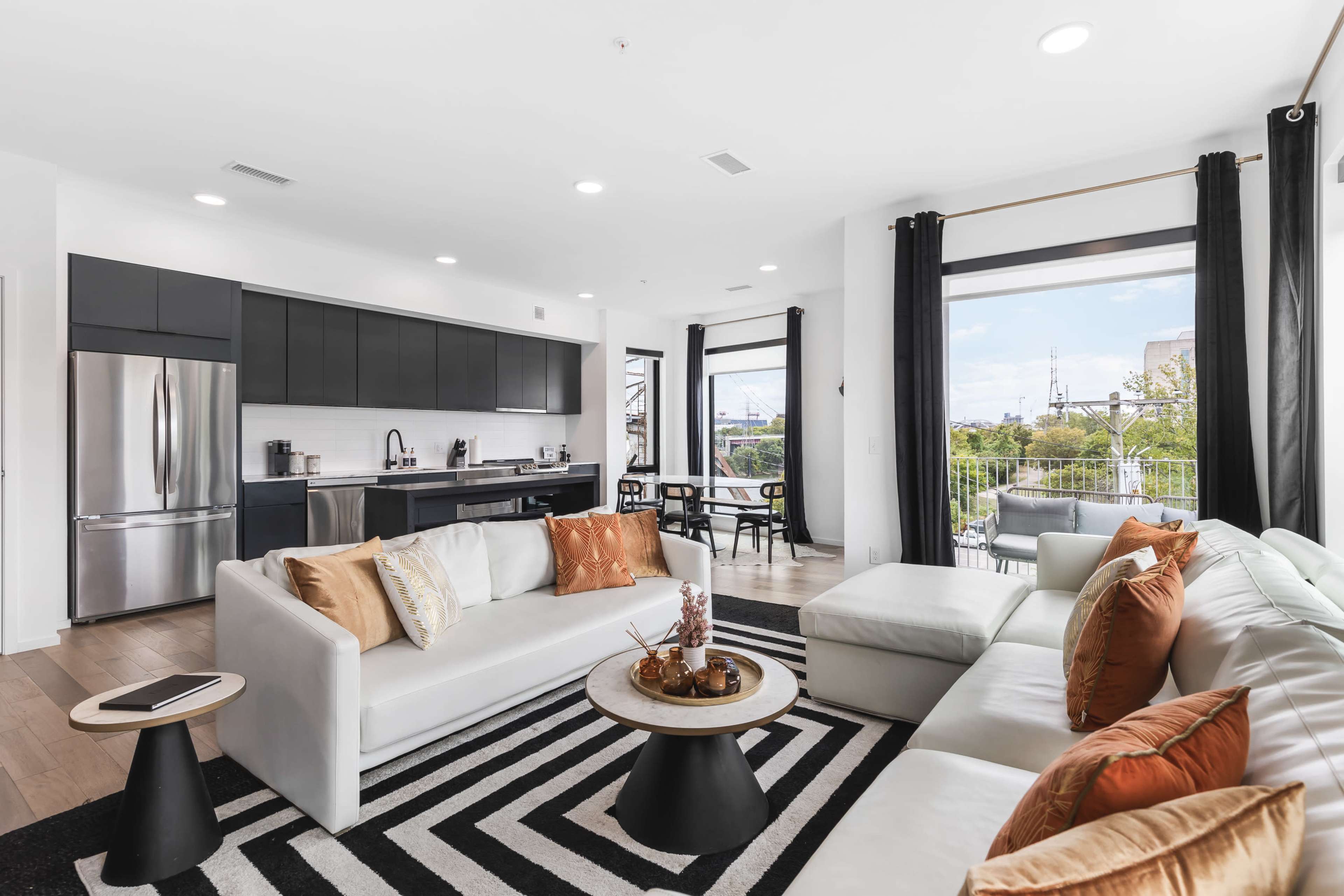 The image shows a modern living room with a sectional sofa, coffee table, and a kitchen area featuring sleek cabinets and a window view.