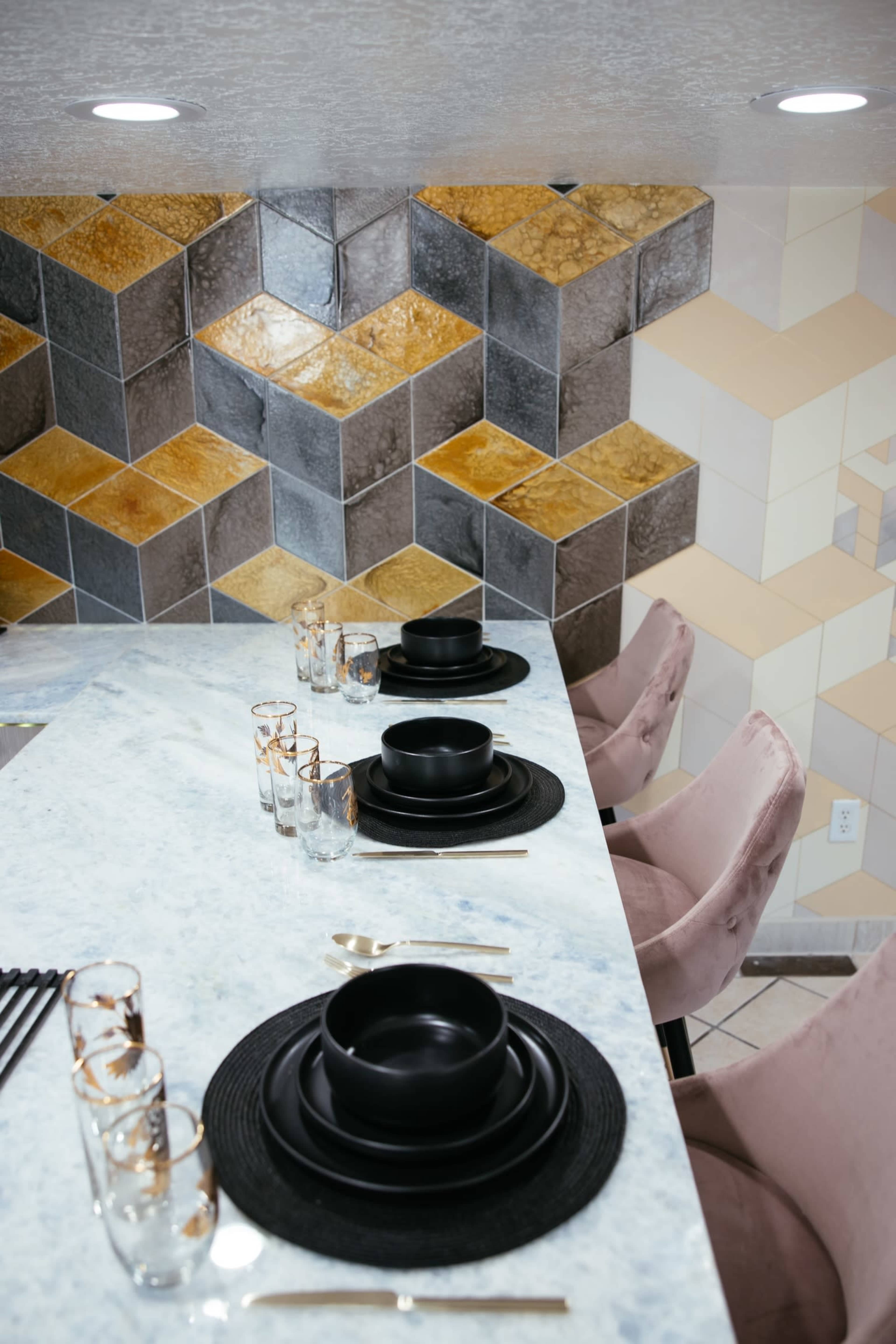 A dining table is set with black plates and bowls on round placemats, alongside clear drinking glasses, against a backdrop of geometric patterned walls in various colors.