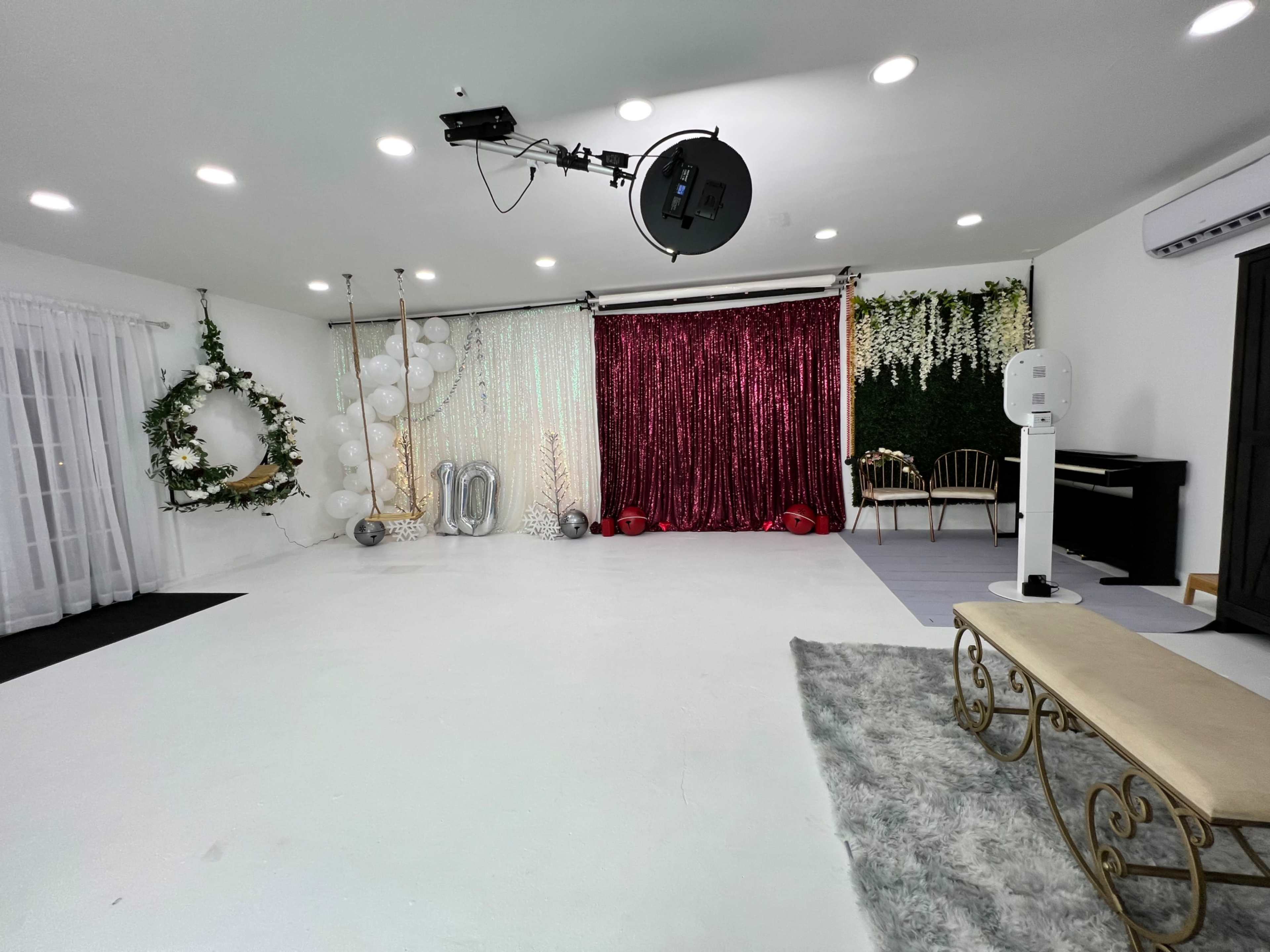 A well-decorated studio space with a white floor, a bench, greenery, balloons, a glittery backdrop, and a piano.