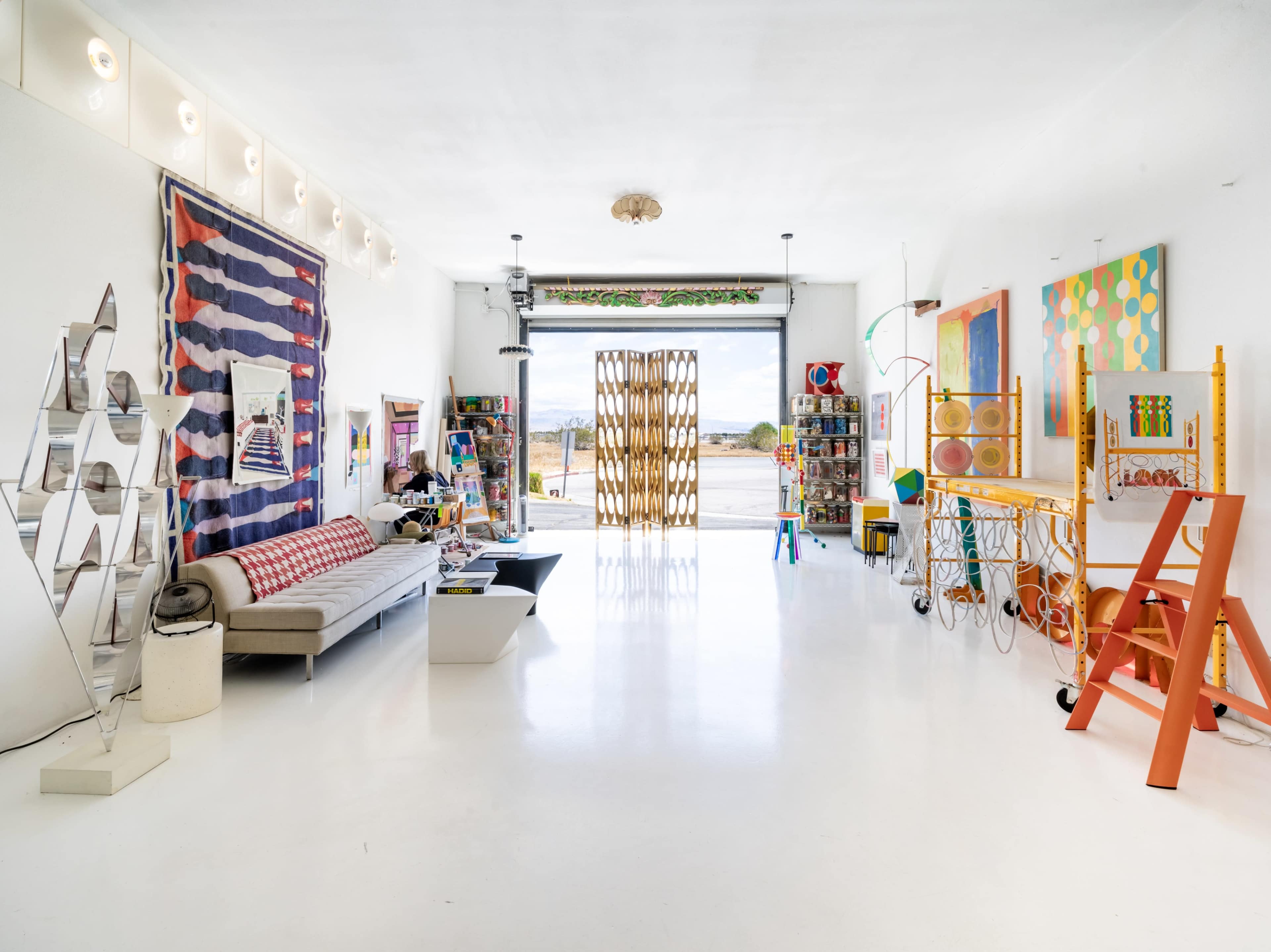 The interior of a brightly lit art studio features colorful artwork, various furniture pieces, and an open entrance showcasing a view outside.