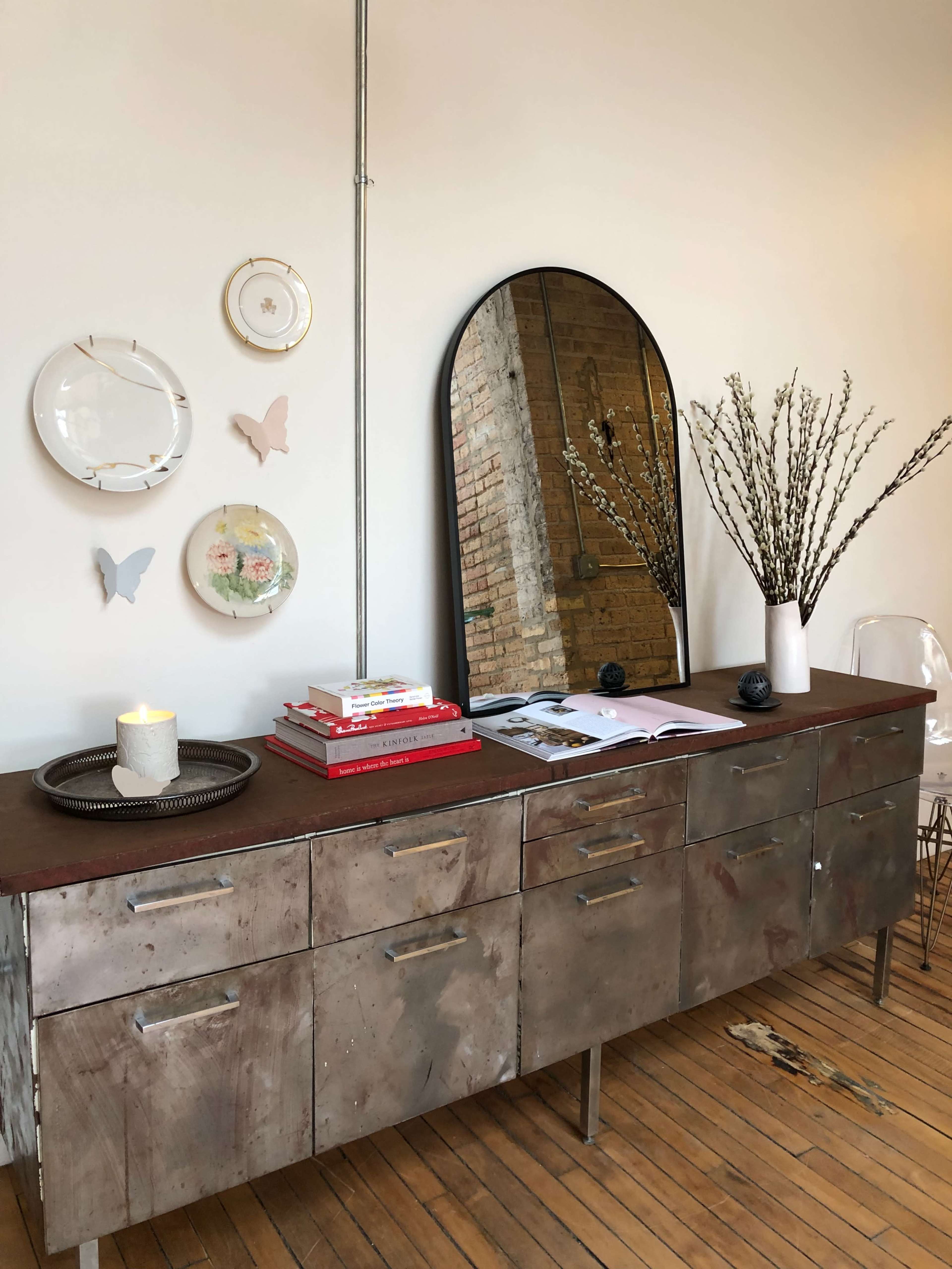 A metal sideboard with several drawers is topped with books, a candle, and a vase of branches, alongside decorative plates and butterflies arranged on the wall.
