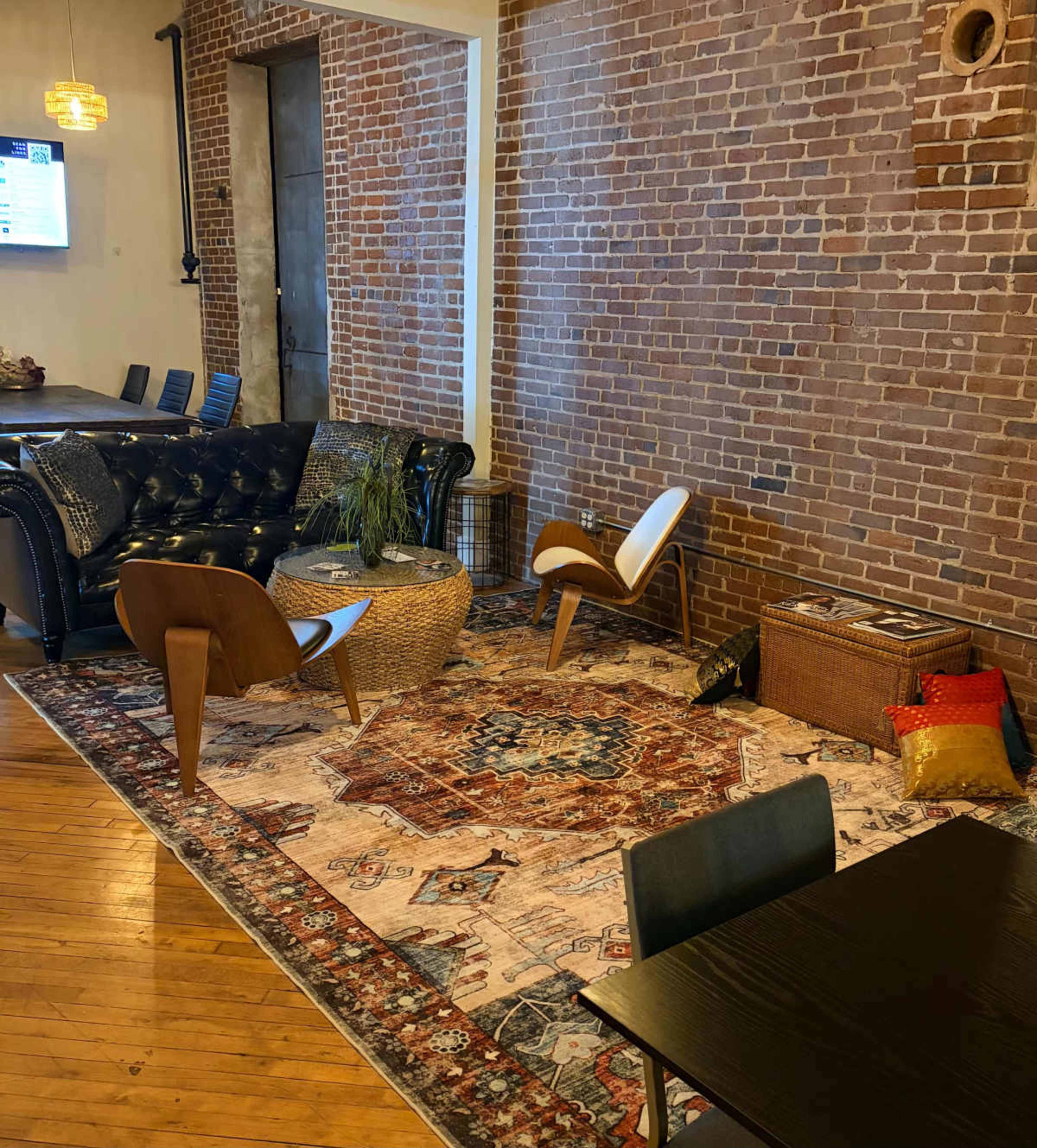 The image shows a cozy sitting area featuring two chairs, a round woven basket, and a colorful area rug set against a backdrop of exposed brick walls.