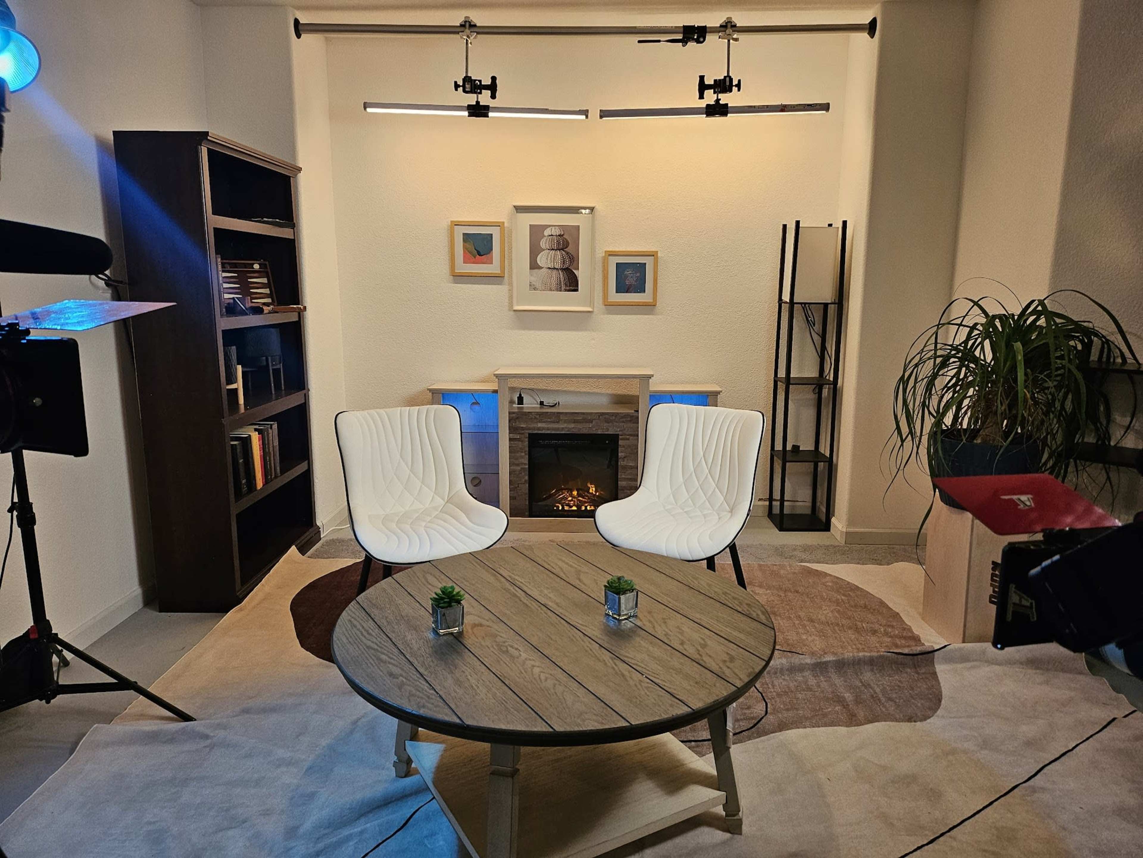 A cozy living room setup features two white chairs facing a small table in front of a fireplace, with bookshelves and a plant in the background.