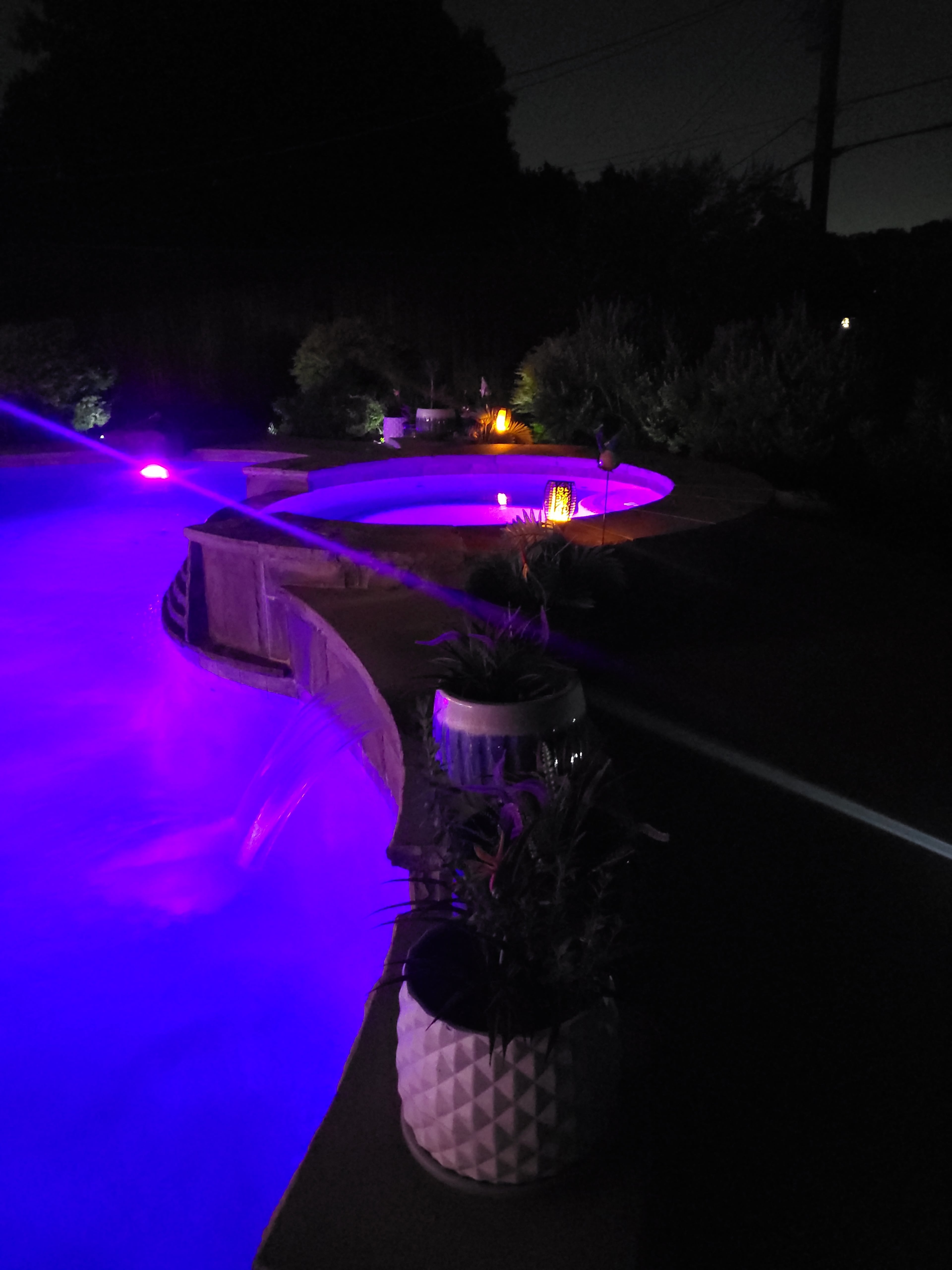 A swimming pool at night, illuminated by colorful lights, with potted plants lining its edge.