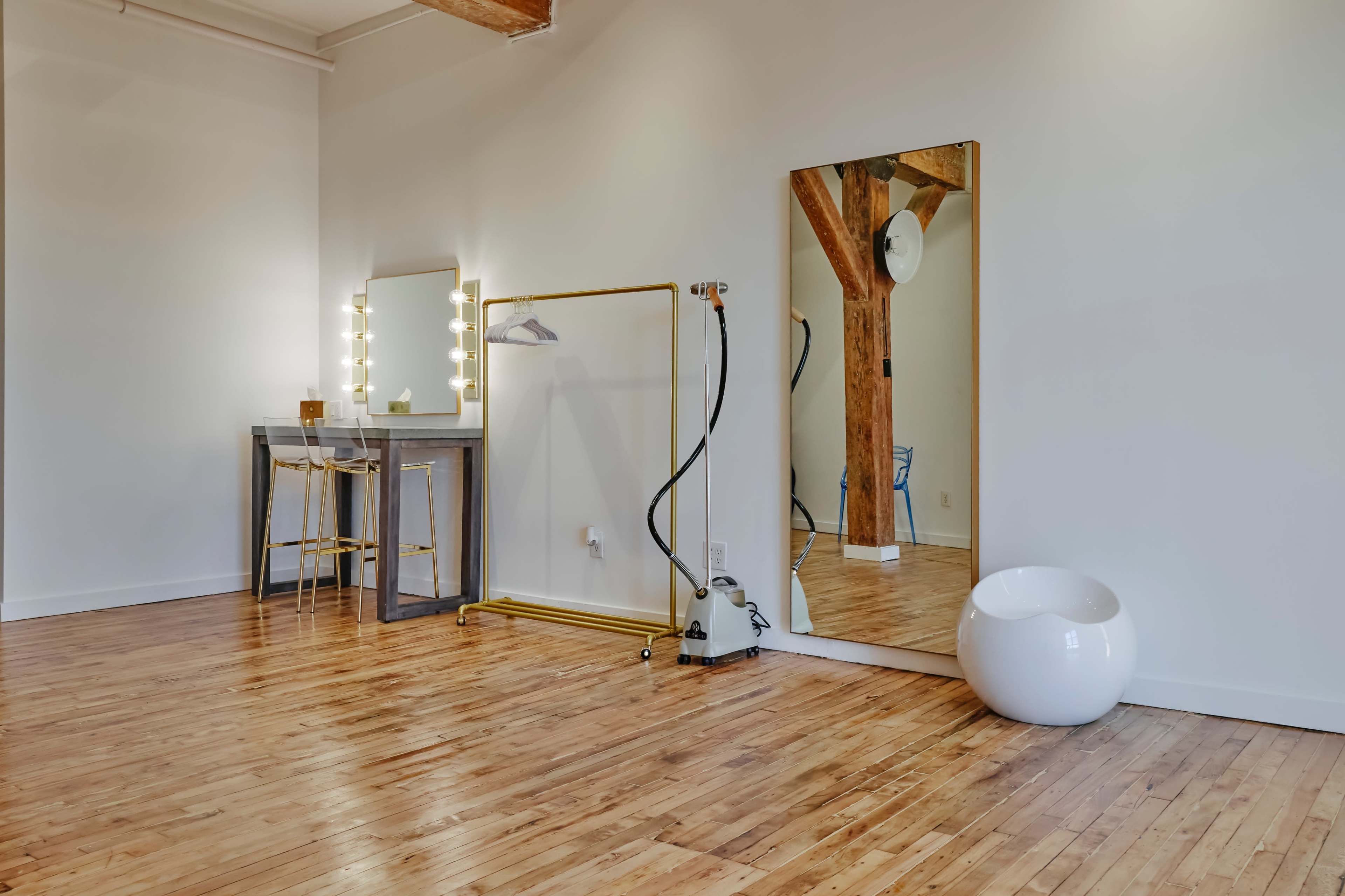 A spacious, well-lit room with wooden floors, featuring a vanity mirror with lights, a clothing rack, a white contemporary chair, and an industrial-style vacuum cleaner.