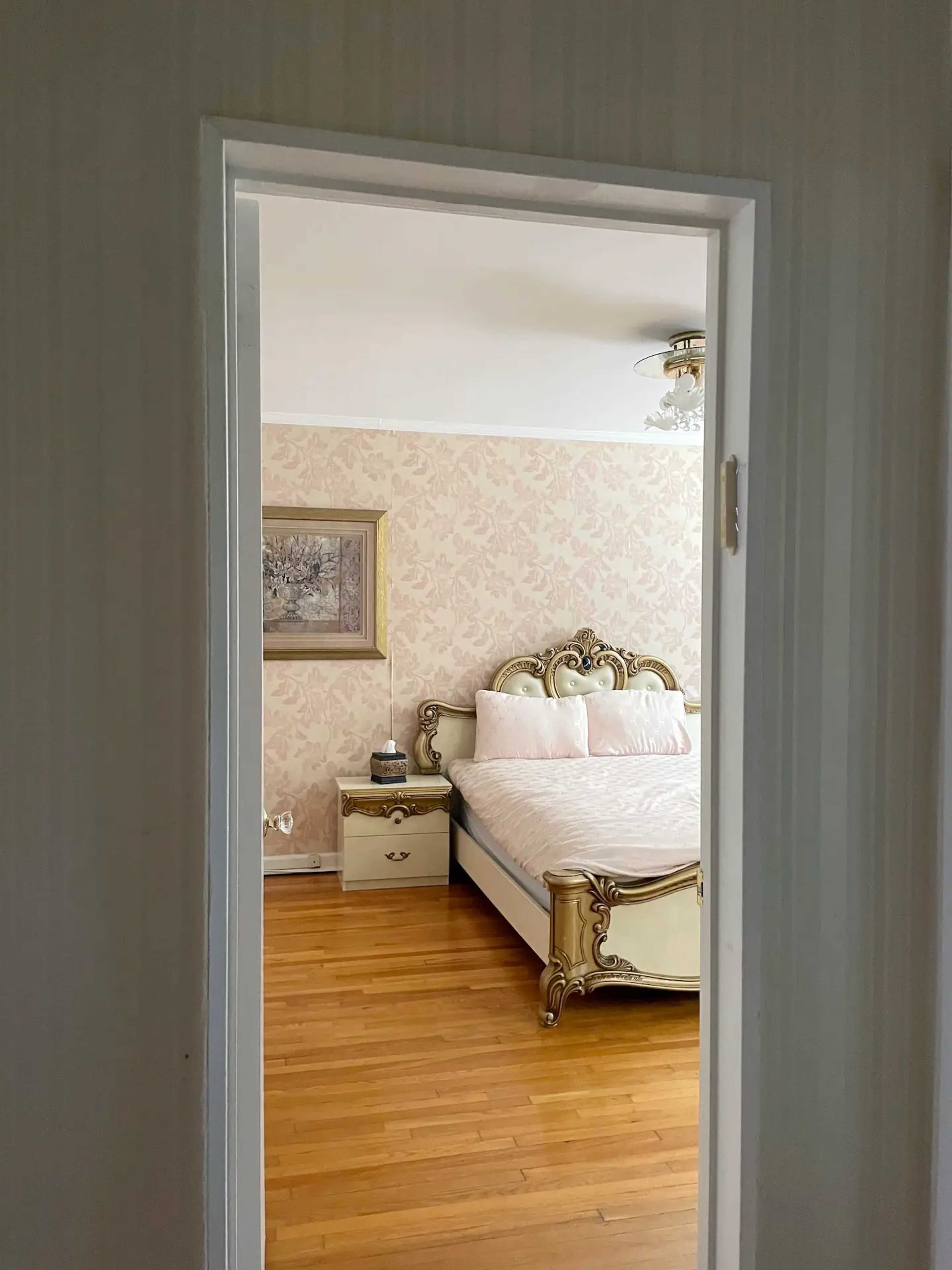 A view into a bedroom with a light-colored wall, a patterned wallpaper, a bed with an ornate headboard, and wooden flooring.