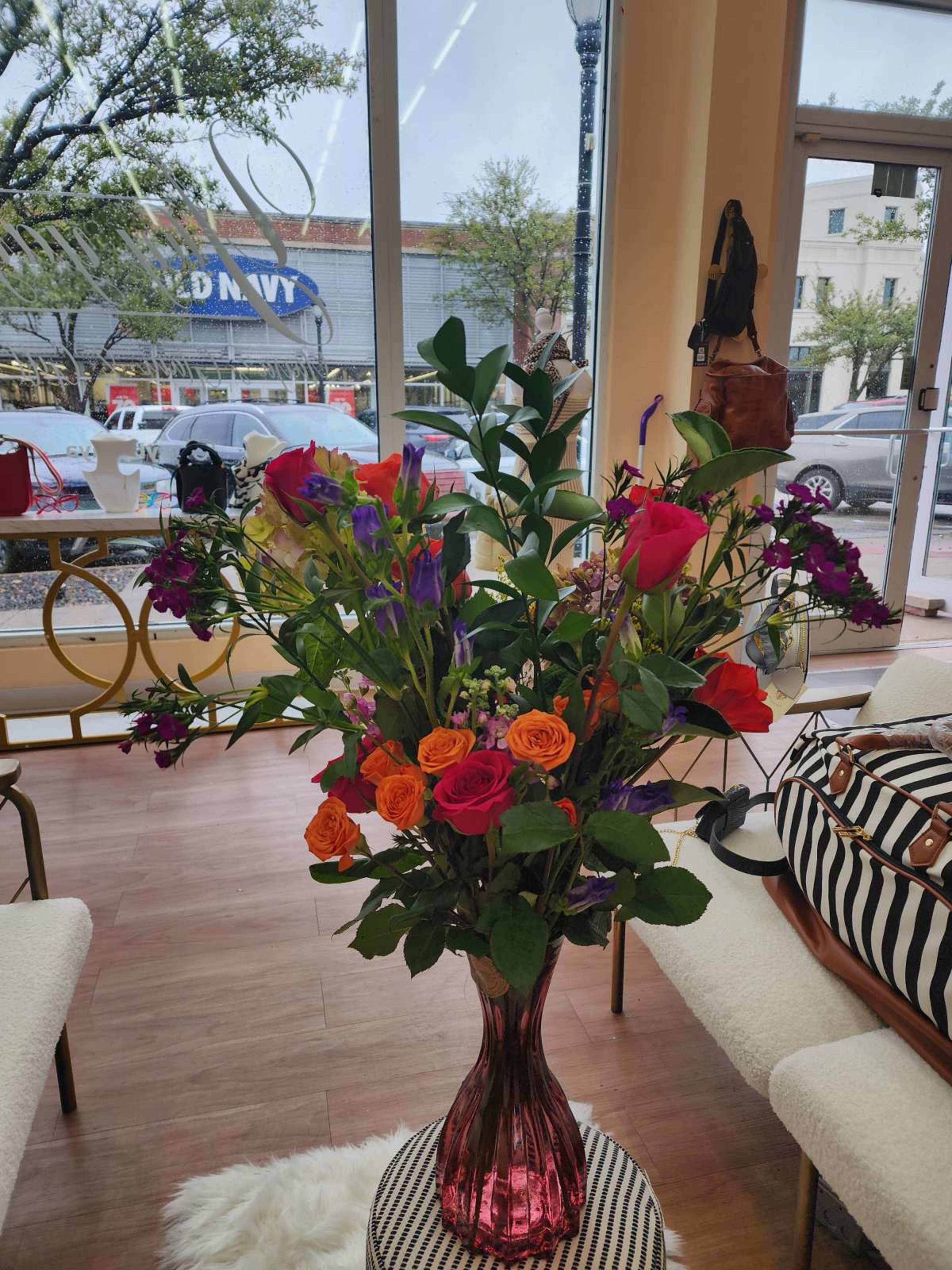 A large bouquet of colorful flowers is displayed in a tall vase on a circular table inside a shop, with a view of parked cars and buildings visible through the window.