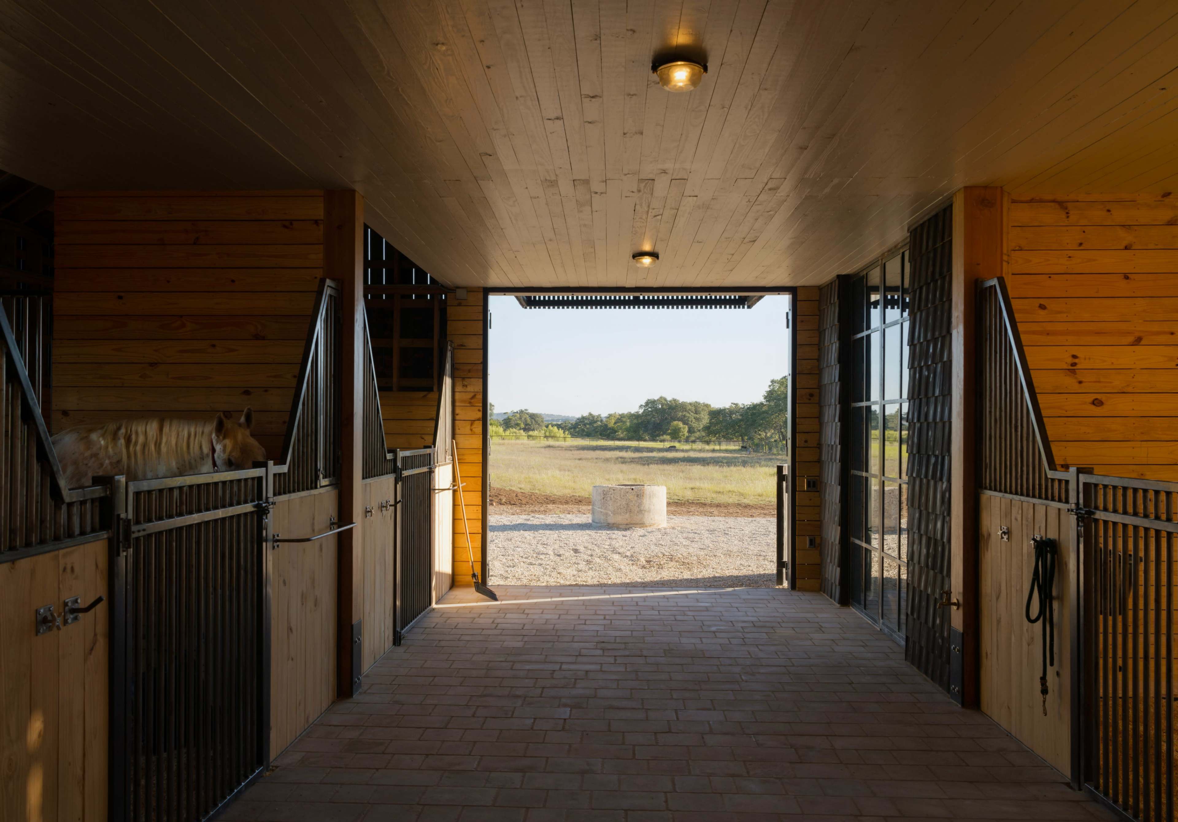 The image shows the interior of a horse stable looking out towards a grassy area through an open entrance.
