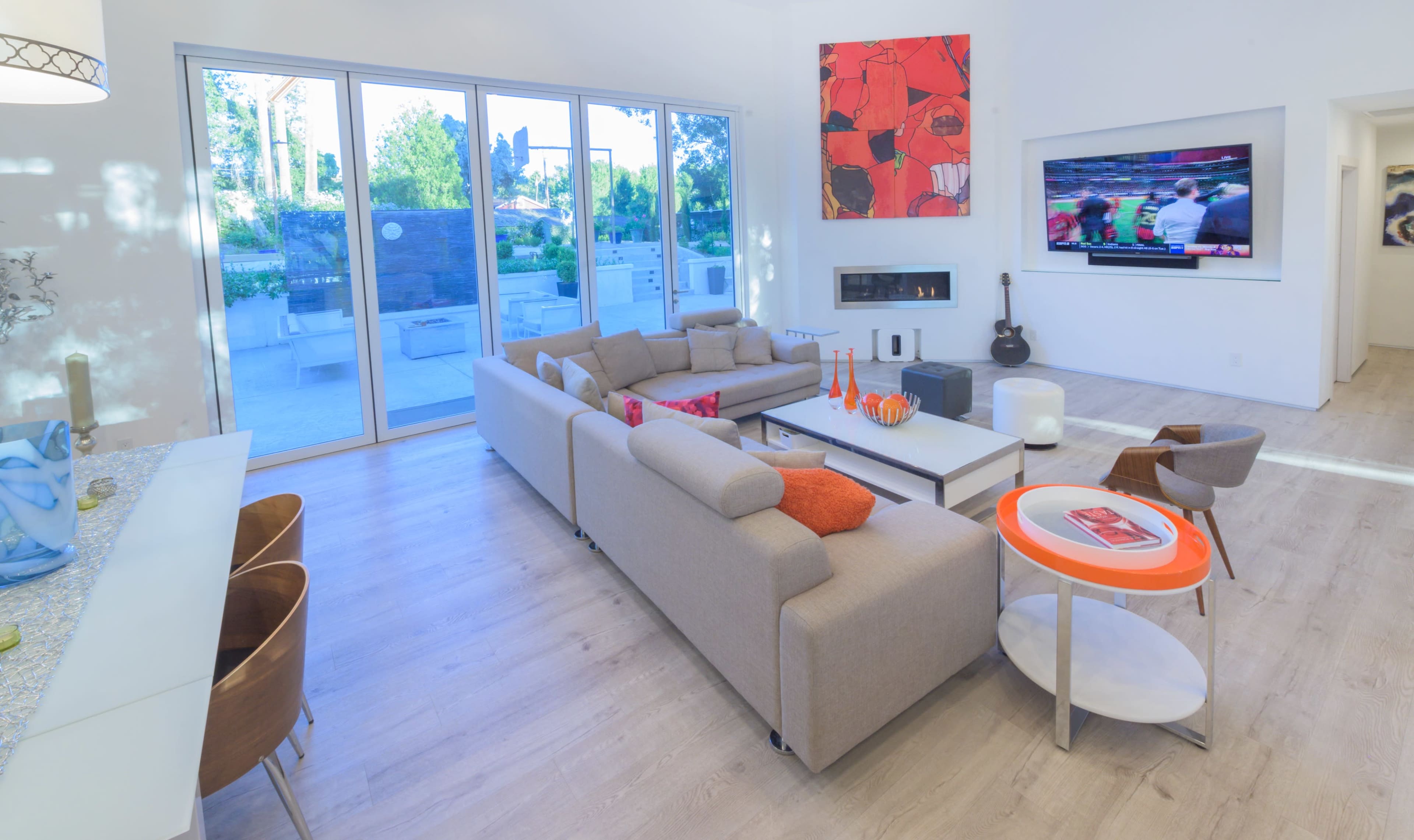 A modern living room features a beige sectional sofa, a television mounted on the wall, and large windows overlooking an outdoor space.