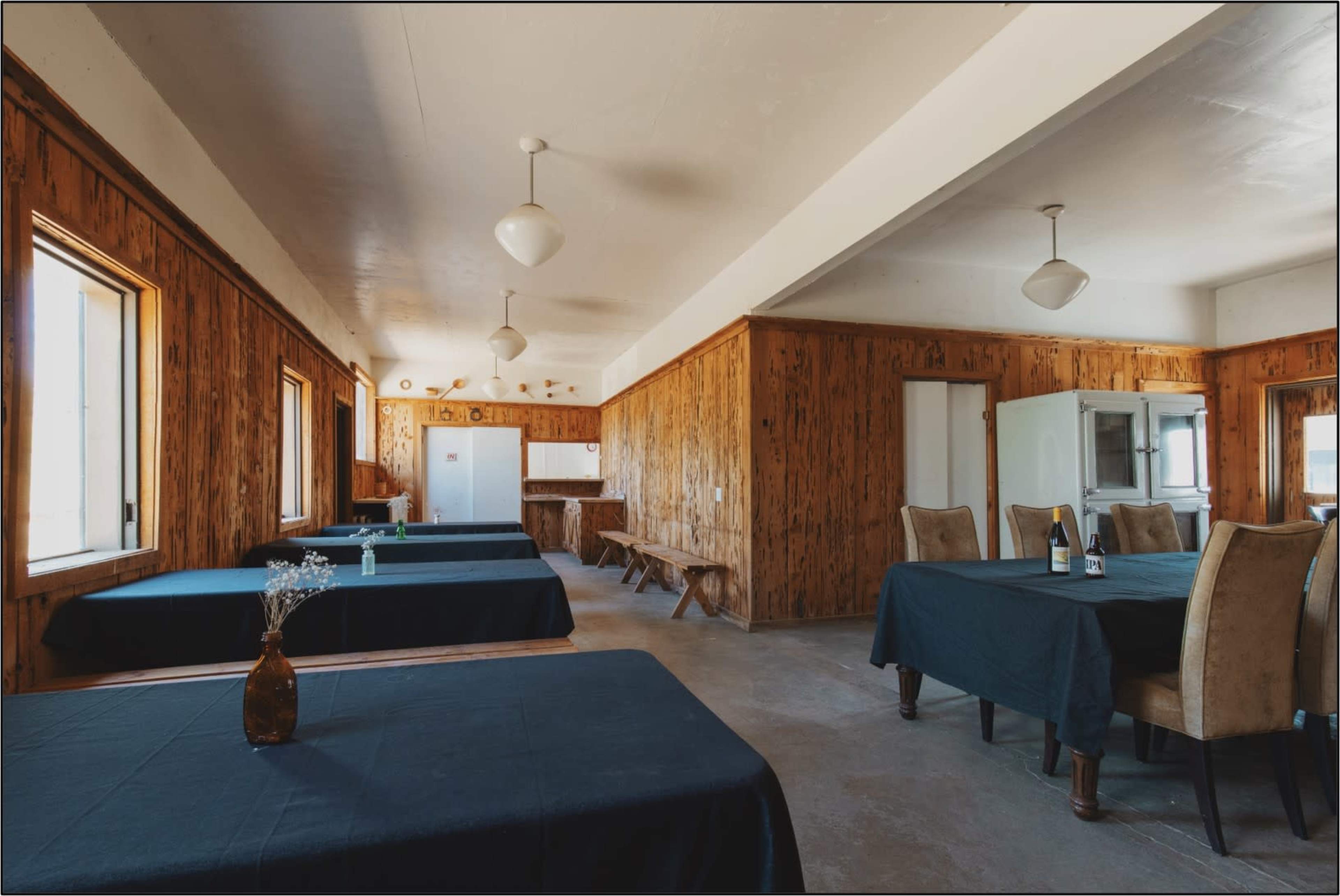 A spacious, sparsely furnished dining area with wooden walls, several tables covered in dark tablecloths, and simple light fixtures hanging from the ceiling.